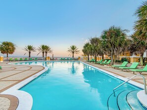 Une piscine entourée d’une clôture blanche, avec des chaises longues vertes et des palmiers.