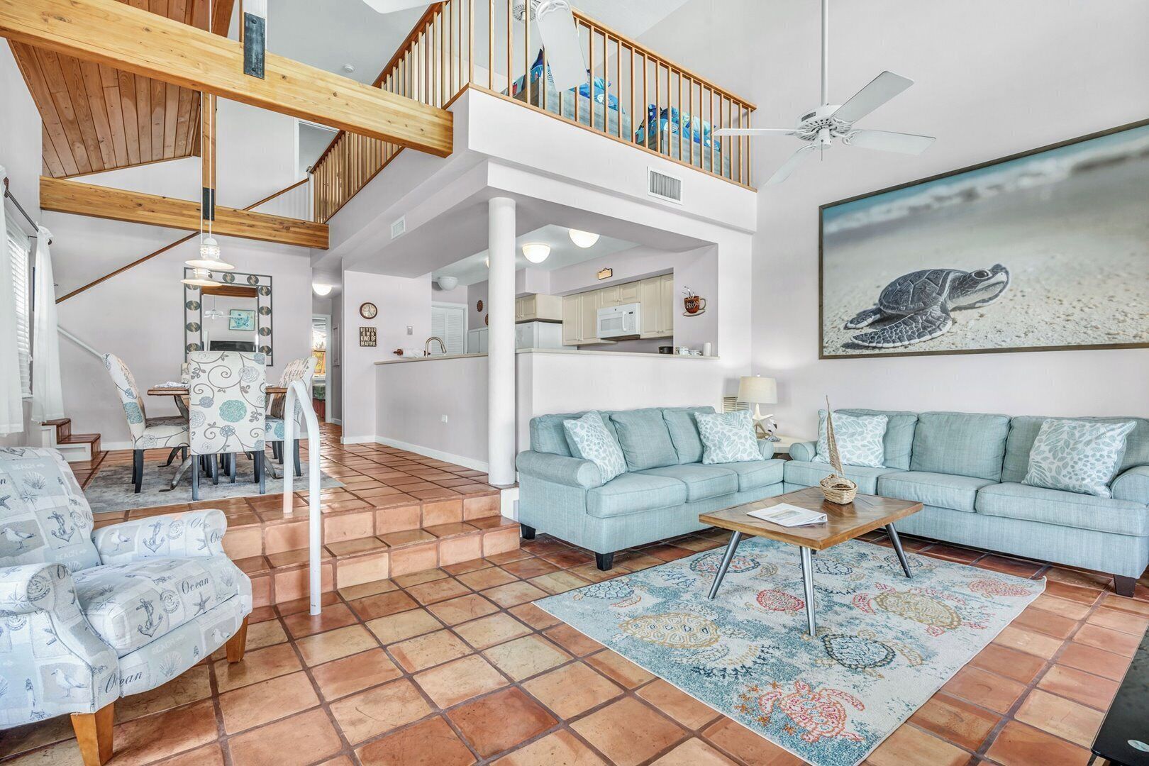 A spacious living area with a high ceiling, a dining area with patterned chairs, a light blue sofa set, and a kitchen in the background.