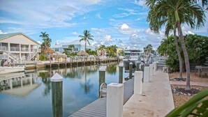 A marina with docked boats, a wooden walkway, and a row of houses with a clear sky. - P63 - Beautiful, waterfront duplex, walking distance to The Cabana Club! (Key Colony Beach)