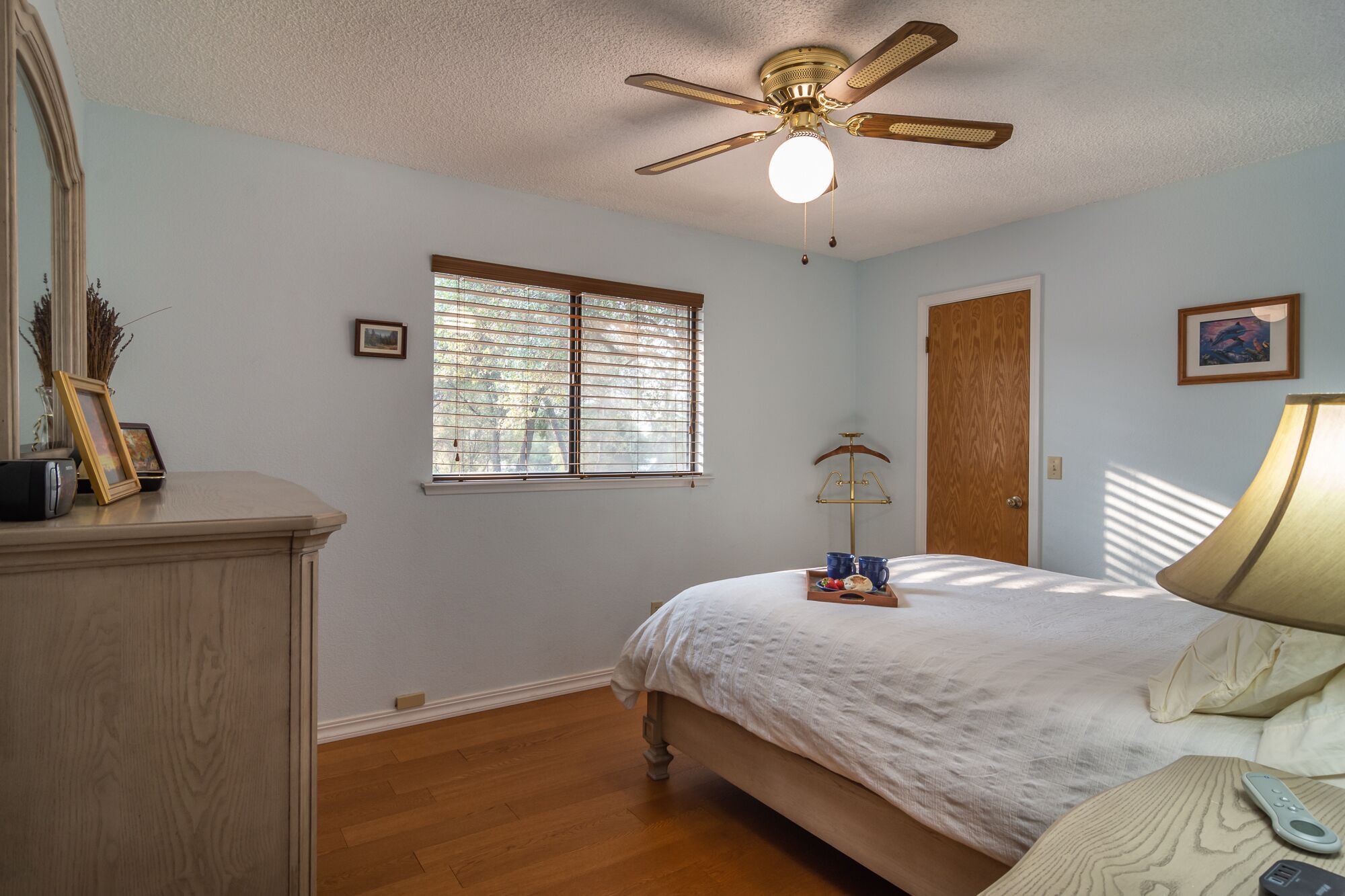 A bedroom with a bed, a ceiling fan, a window with blinds, a wooden door, and a dresser.