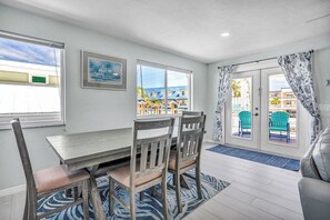 A dining area with a wooden table and chairs, a blue patterned rug, and a view of a building with palm trees through the window. - M123 - Cabana Beach Duplex! Premier location across the street from Cabana Beach Club! (Key Colony Beach)