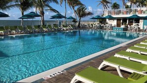 Une piscine à l’eau d’un bleu cristallin, entourée de chaises longues et de parasols, avec des palmiers et un bâtiment en arrière-plan.