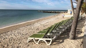 Une plage bordée de rangées de chaises longues vertes et blanches, un brise-lames rocheux et un ciel bleu dégagé.