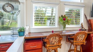 Une cuisine avec un plan de travail en bois, deux chaises en osier, un vase de fleurs et une vue sur le jardin par la fenêtre.