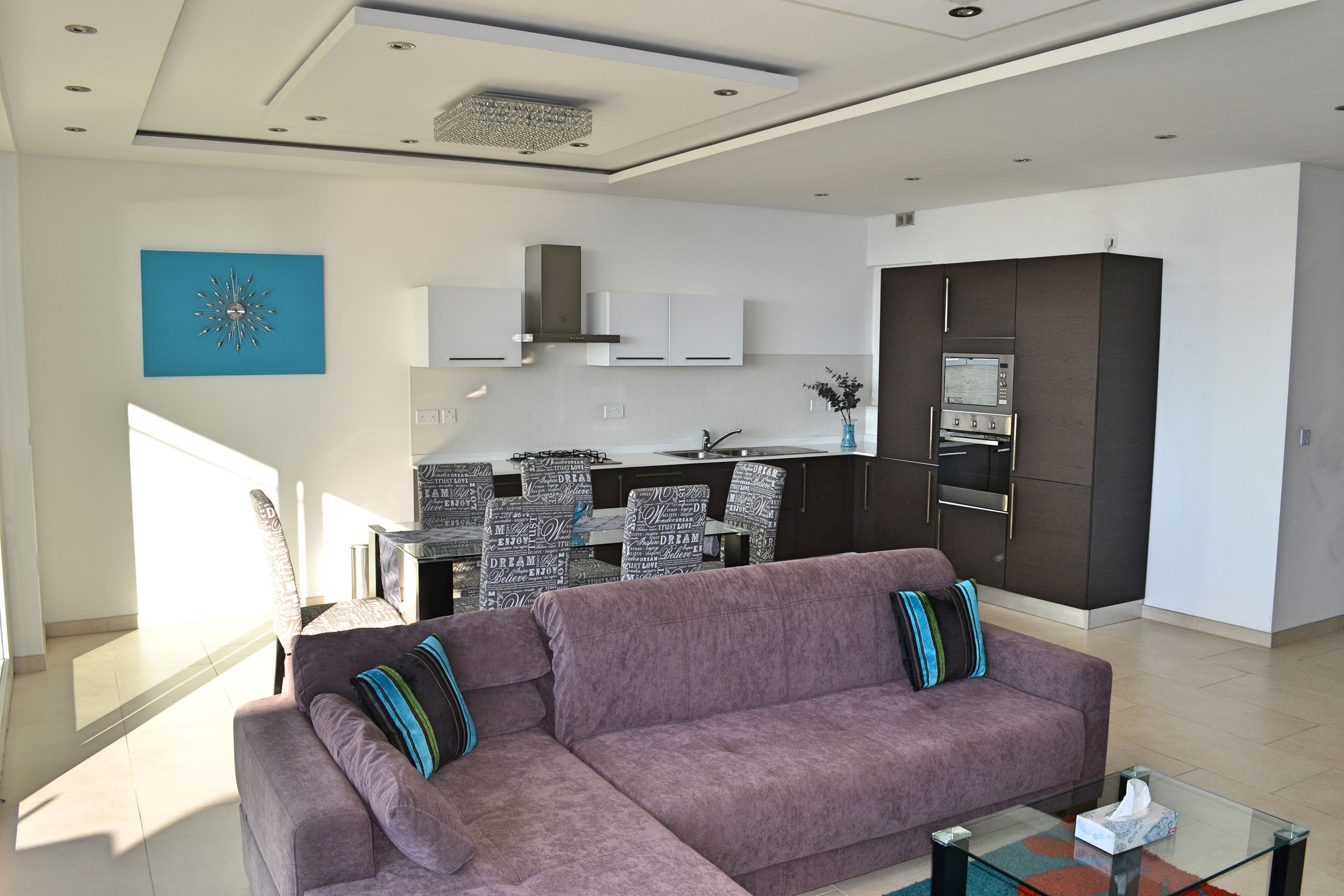 A modern living room with a sofa, a glass coffee table, and a kitchen area in the background.