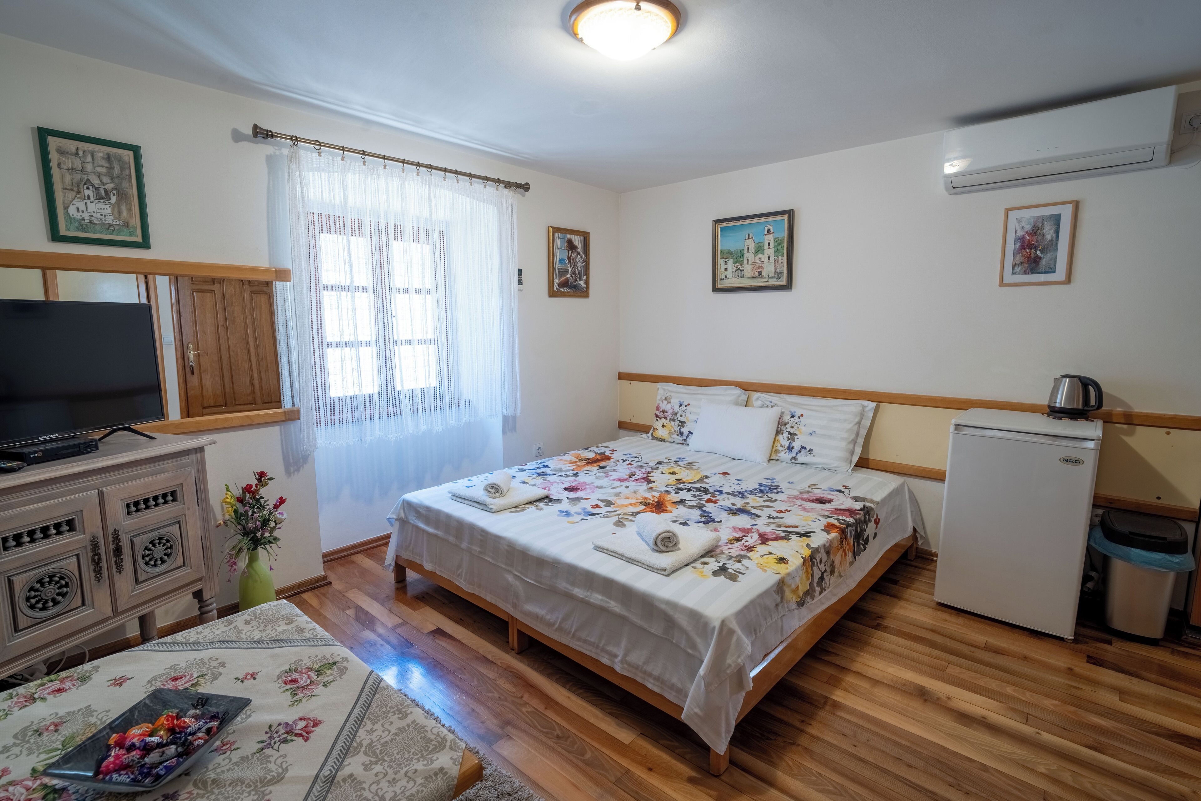 A bedroom with a wooden bed, a television on a cabinet, a refrigerator, and a window with curtains.