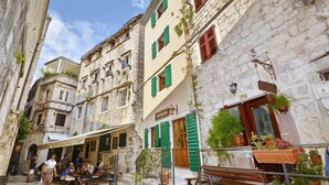 A narrow street with buildings featuring stone facades and green shutters, a café with outdoor seating, and potted plants. - Studios Belvi (Kotor)