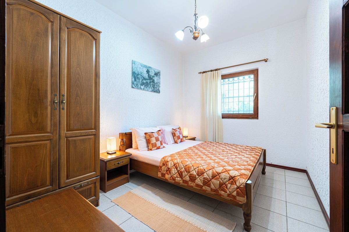 A bedroom with a wooden wardrobe, a bed with a quilt, a nightstand, a bedside table, and a window with curtains.