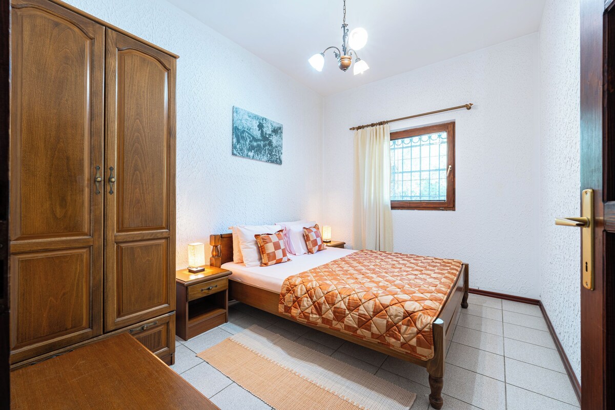 A bedroom with a wooden wardrobe, a bed with a quilt, a nightstand, a bedside table, and a window with curtains.