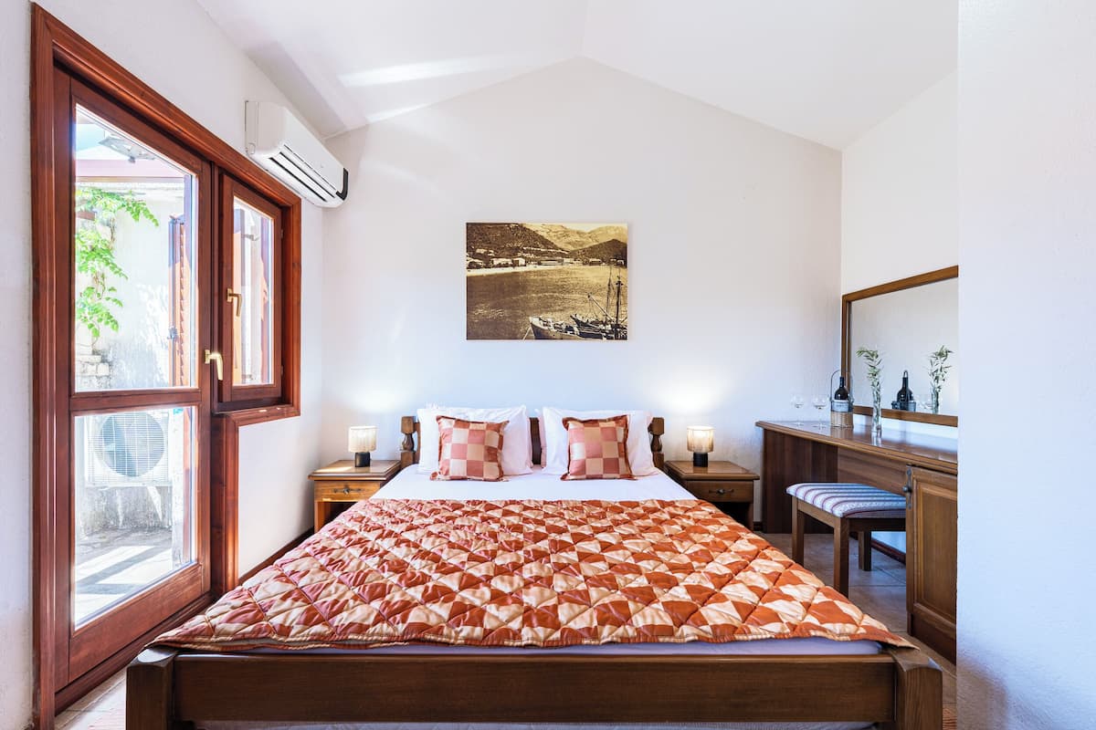 A bedroom with a bed, a bedside table with a lamp, a window with an air conditioner, and a painting on the wall.