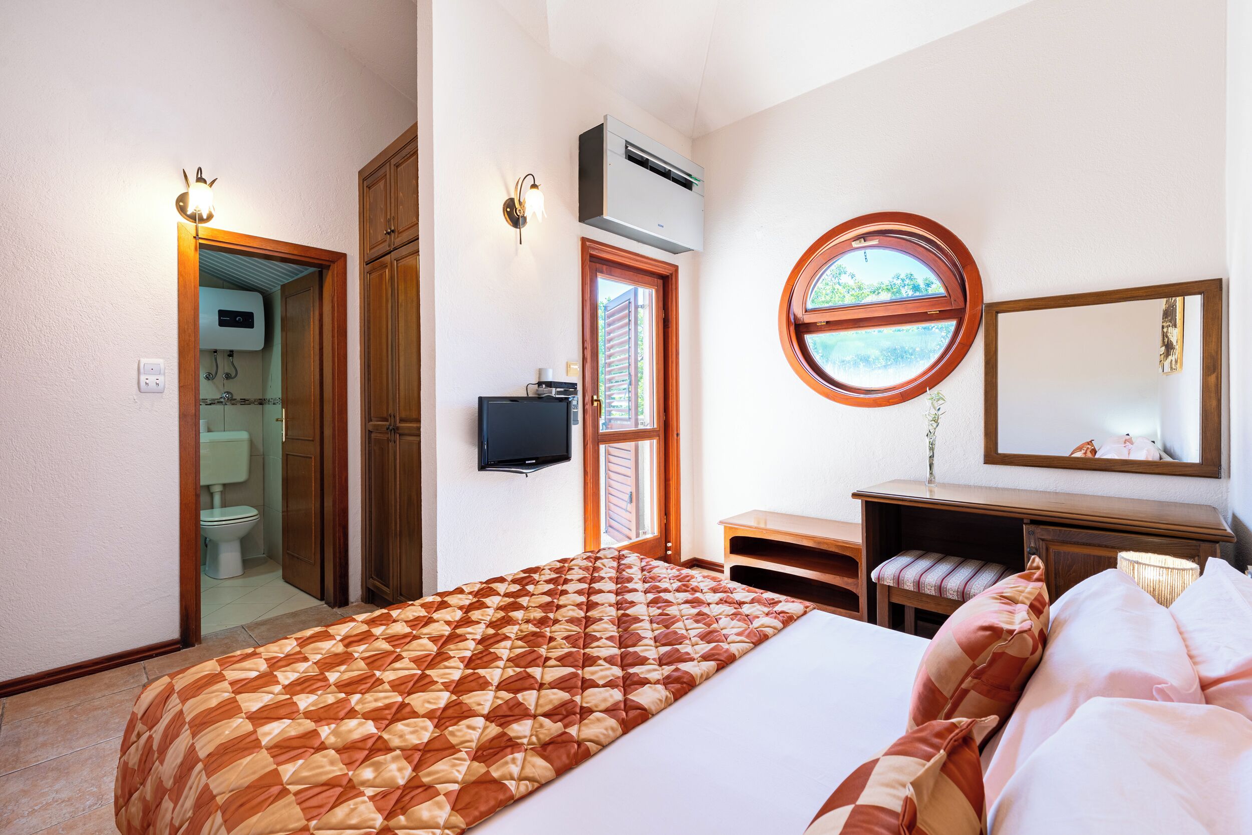 A hotel room with a bed, a desk, a chair, a television, and an air conditioning unit.
