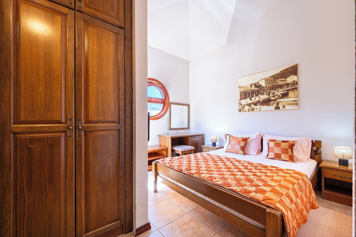 Quarto com cama, guarda-roupa de madeira, mesa de cabeceira, abajur e um quadro na parede.