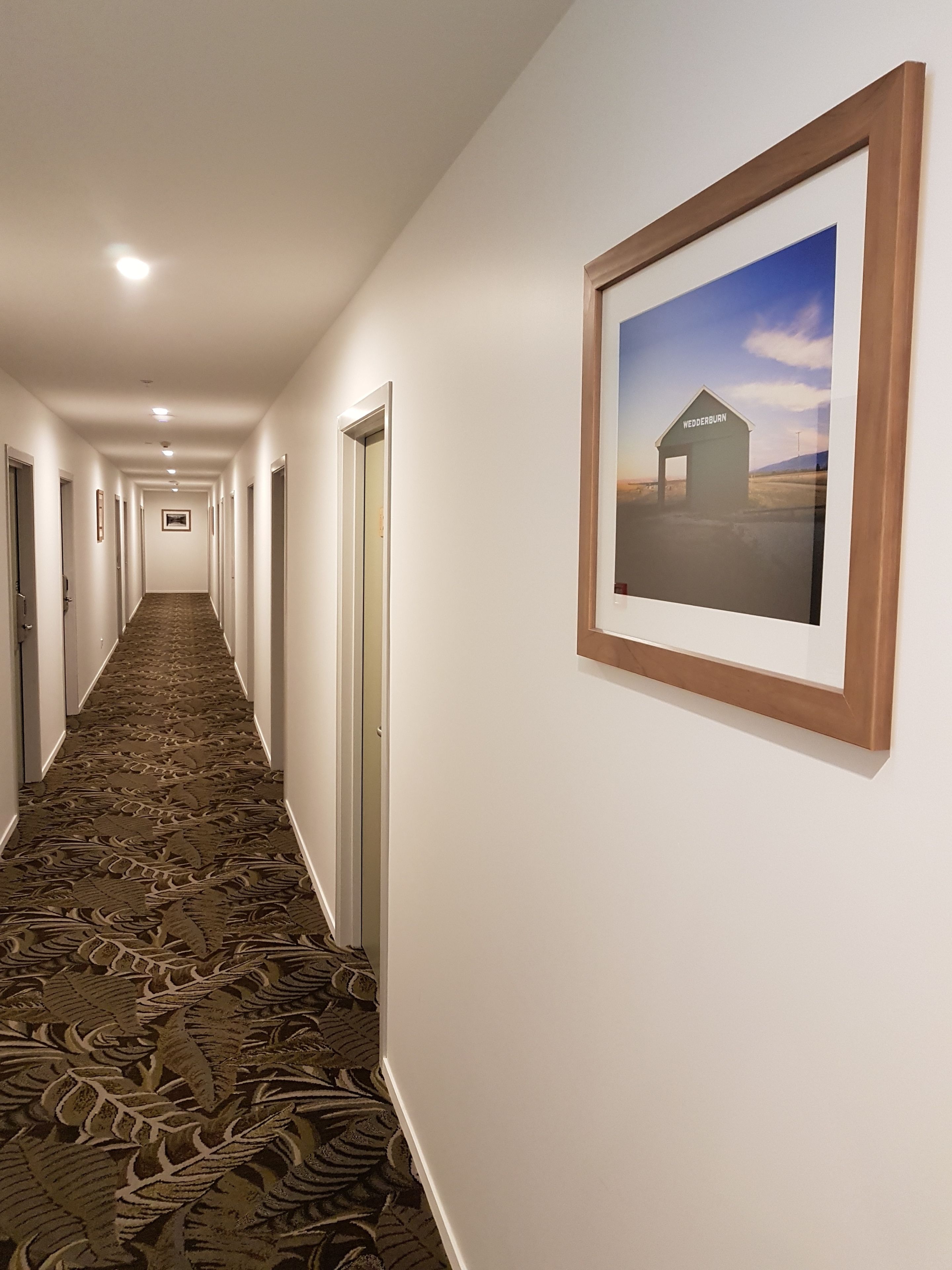 A hotel hallway with a patterned carpet, white walls, and framed pictures on the wall.