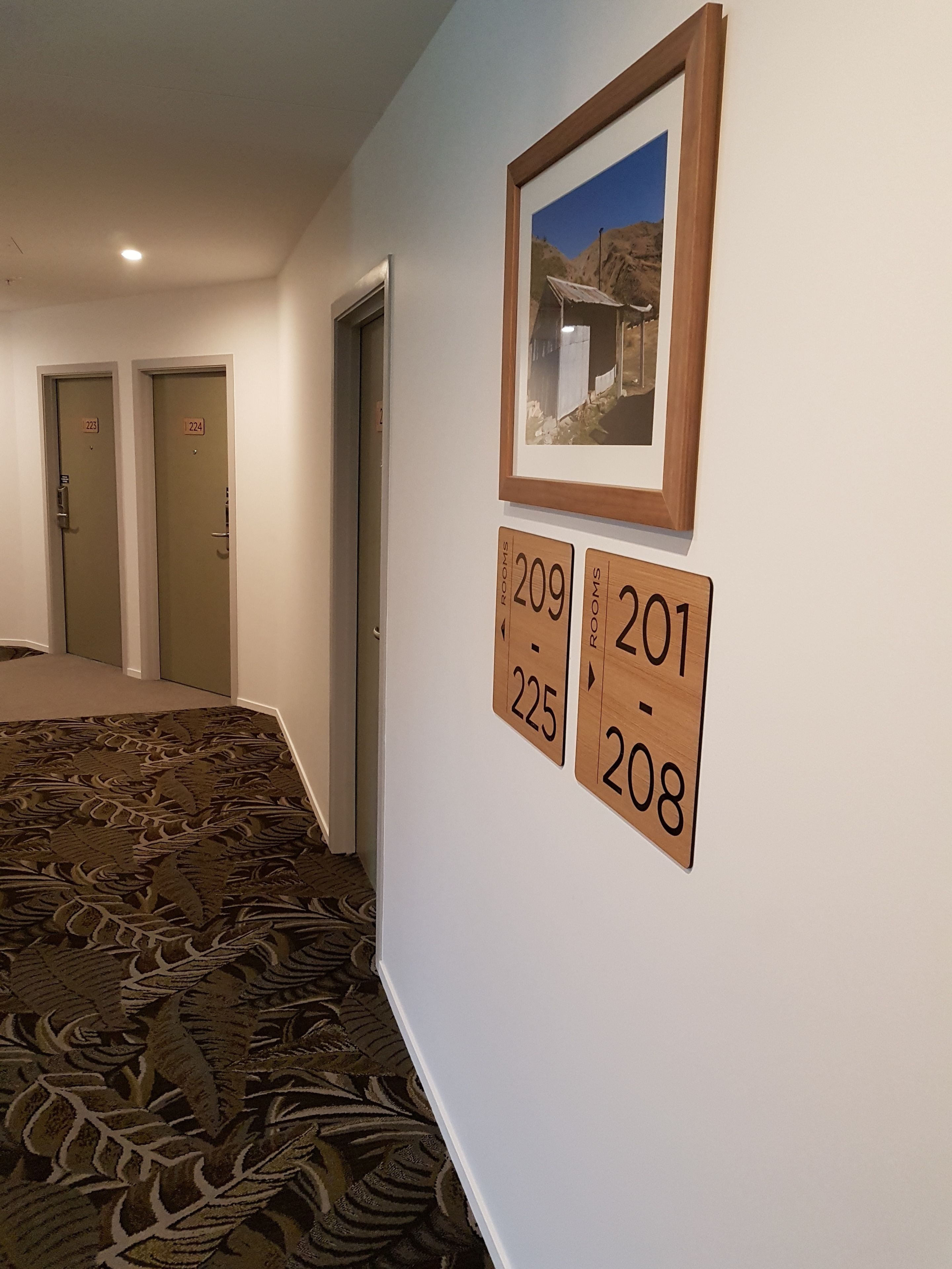 Un pasillo de hotel con letreros de habitaciones numeradas, un cuadro enmarcado en la pared y suelo alfombrado.
