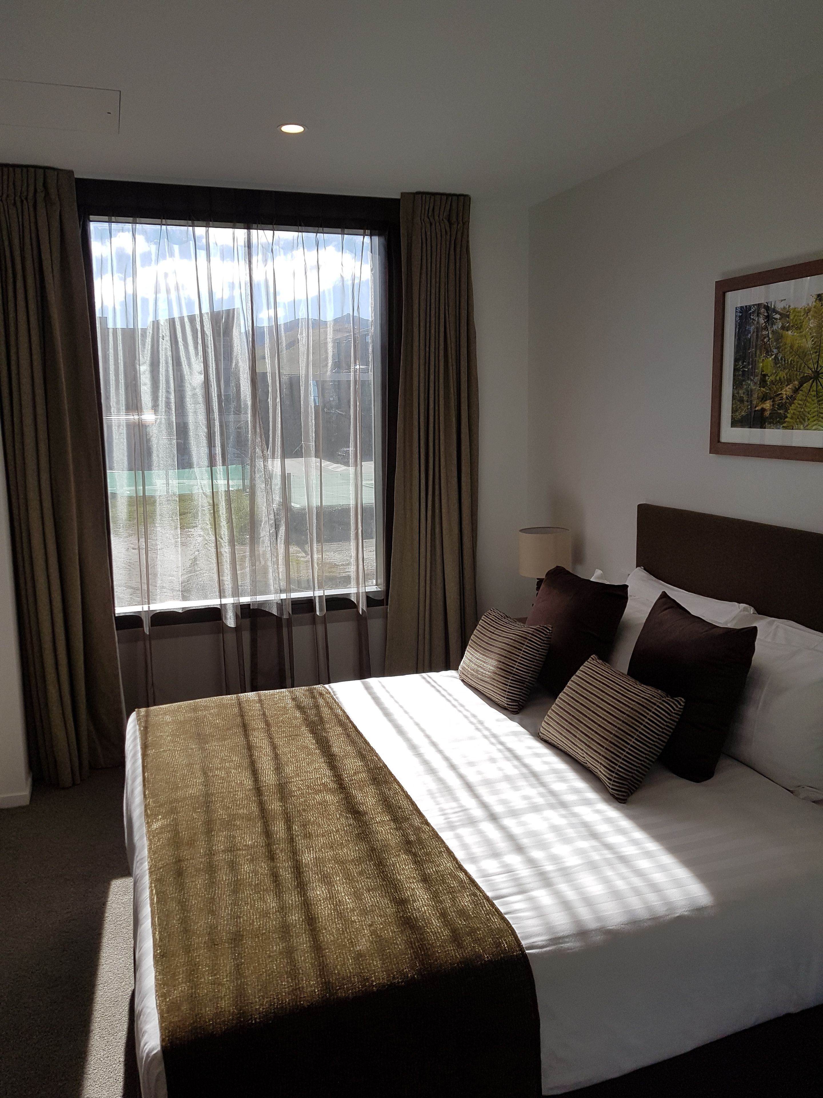 Una habitación de hotel con una cama grande, cabecera, ventana con cortinas e imagen enmarcada en la pared.