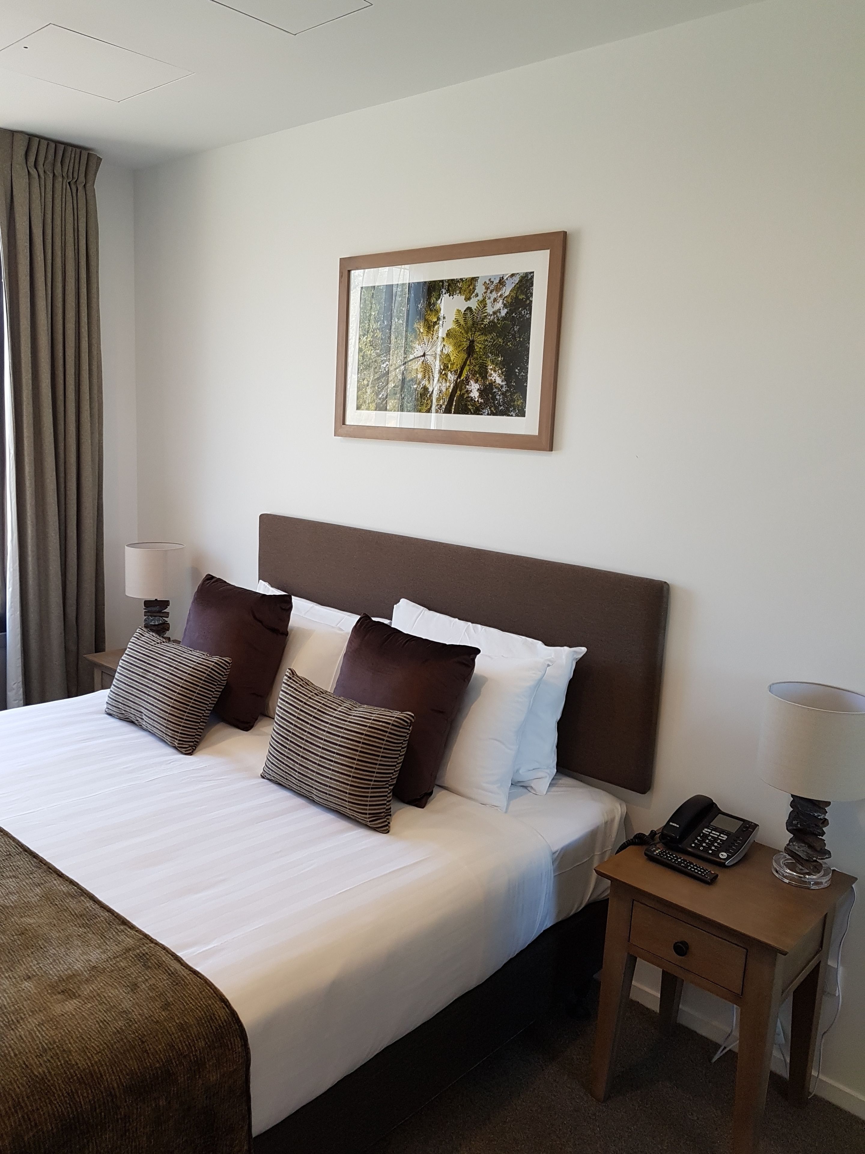 A hotel room with a large bed, a headboard, a bedside table with a phone, a lamp, and a framed picture on the wall.