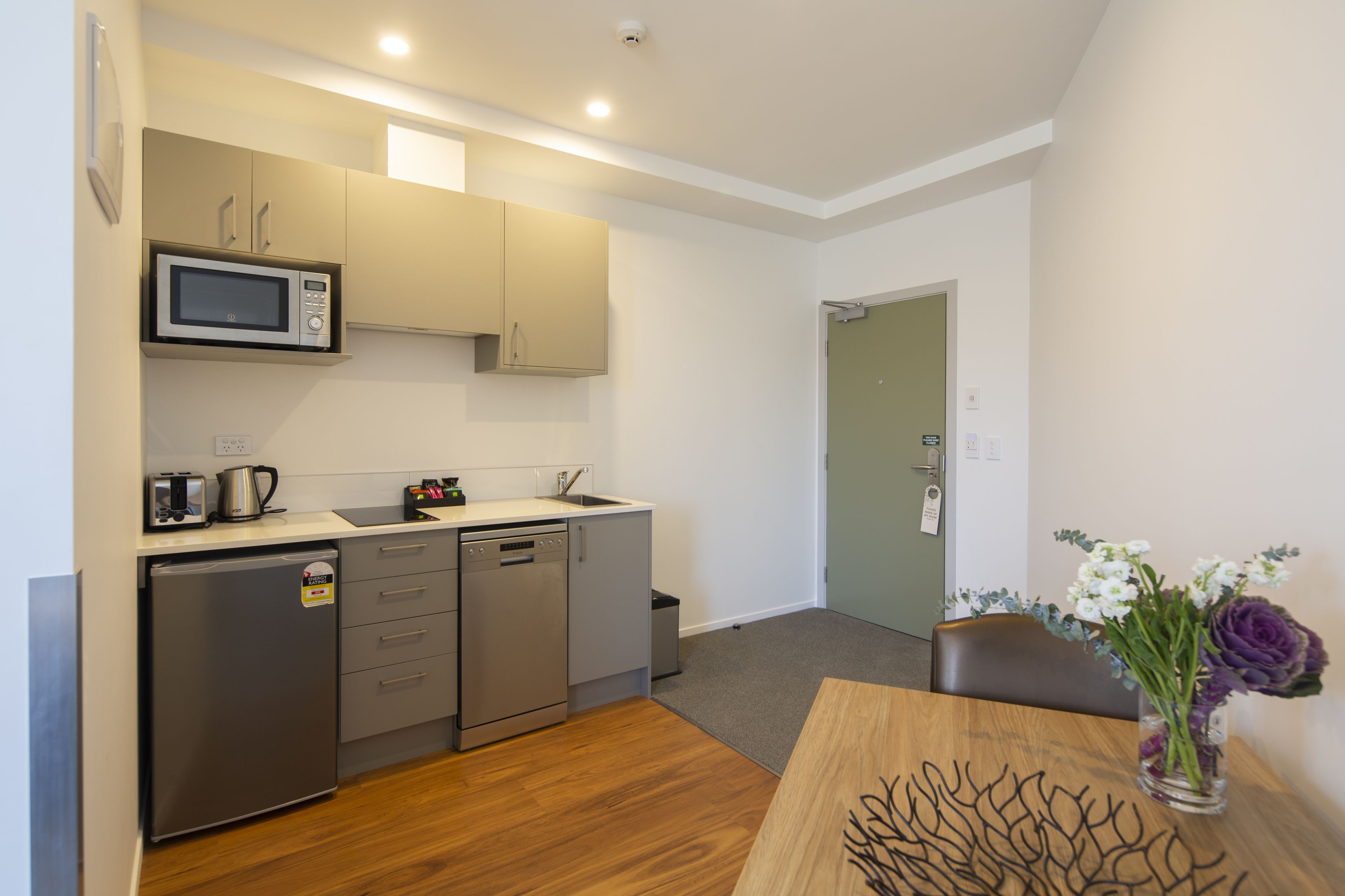 A compact kitchen with a microwave, toaster, and dishwasher. There's a small trash bin and a wooden table with a vase of flowers.