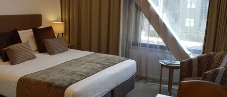 A hotel room with a bed, a wooden table, a small side table, and a view of buildings through the window.