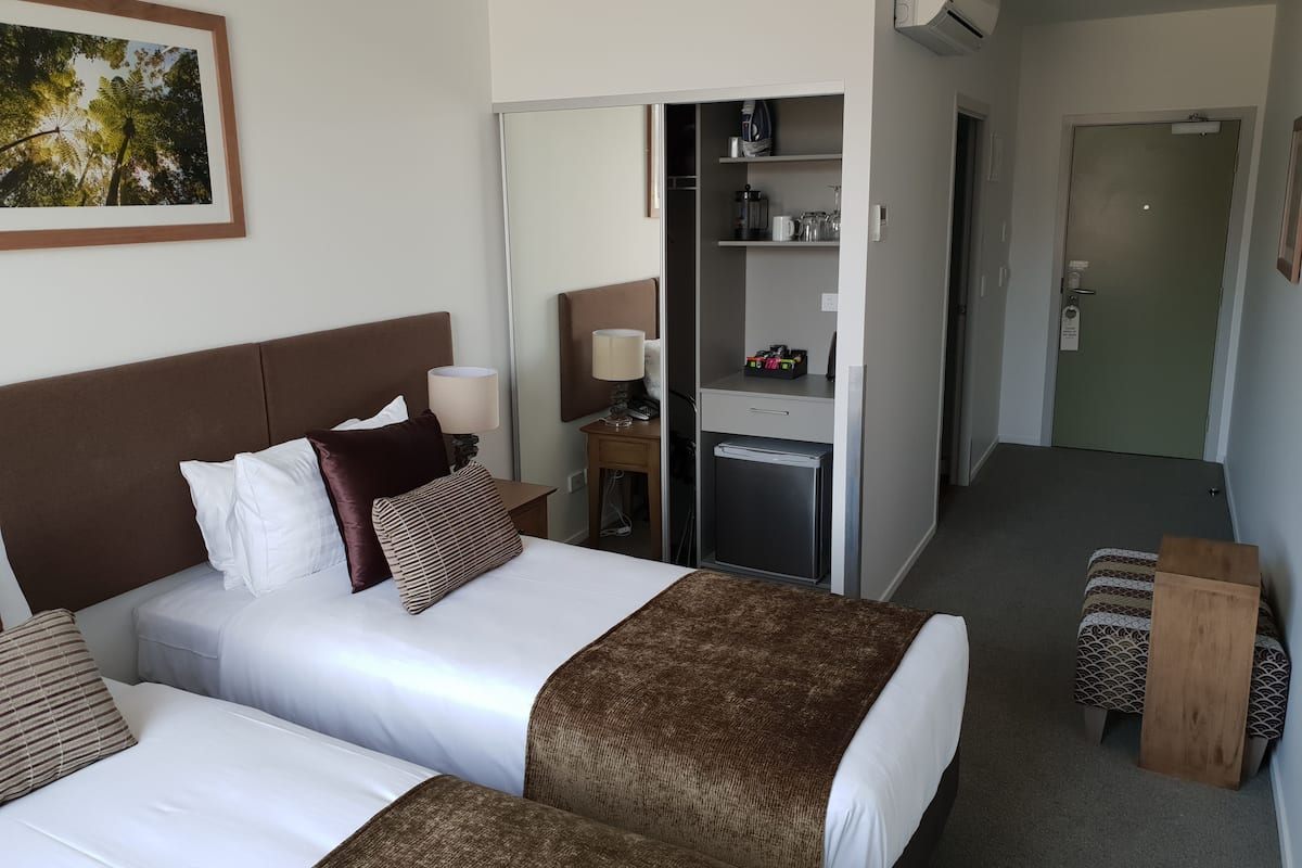 A hotel room with two beds, a wardrobe, a small table with a lamp, and a refrigerator.