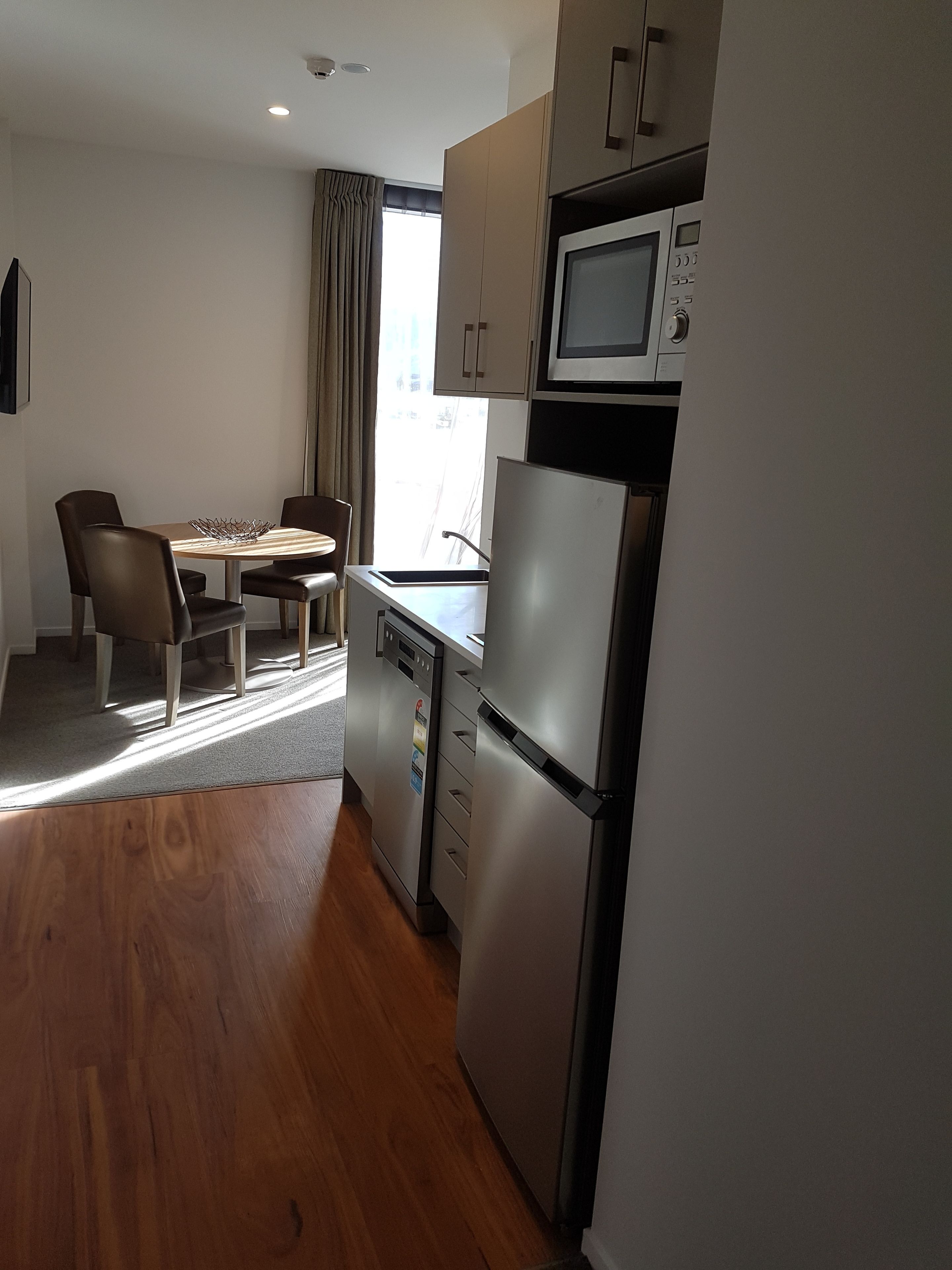 A compact kitchen with a refrigerator, microwave, and dishwasher. There is a dining table with chairs and a window with curtains.