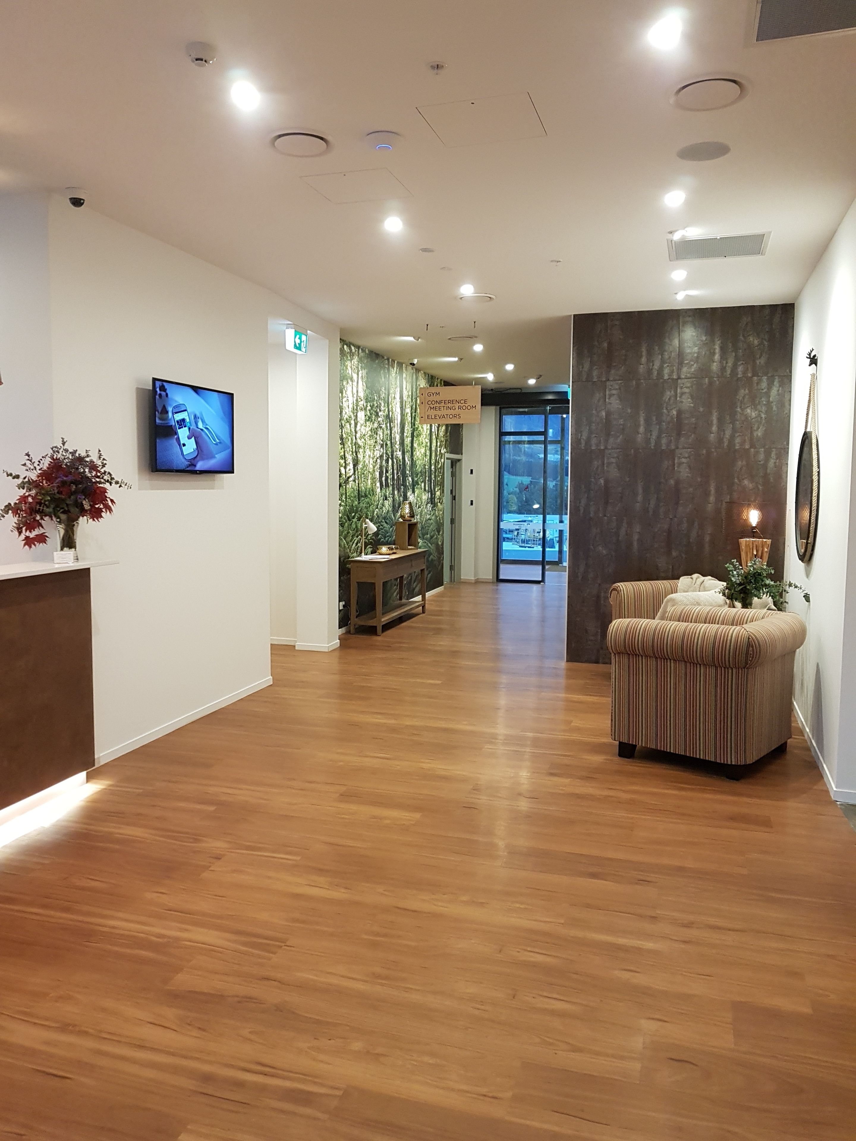 Een hotel lobby met een houten vloer, een receptiebalie, een televisie, een bord dat een sportschool, vergaderruimte en liften aangeeft, en een zithoek met een plant.