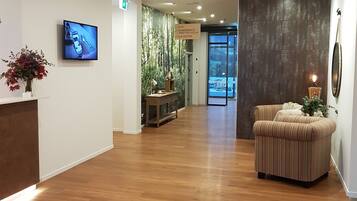 Een hotel lobby met een houten vloer, een receptiebalie, een televisie, een bord dat een sportschool, vergaderruimte en liften aangeeft, en een zithoek met een plant.