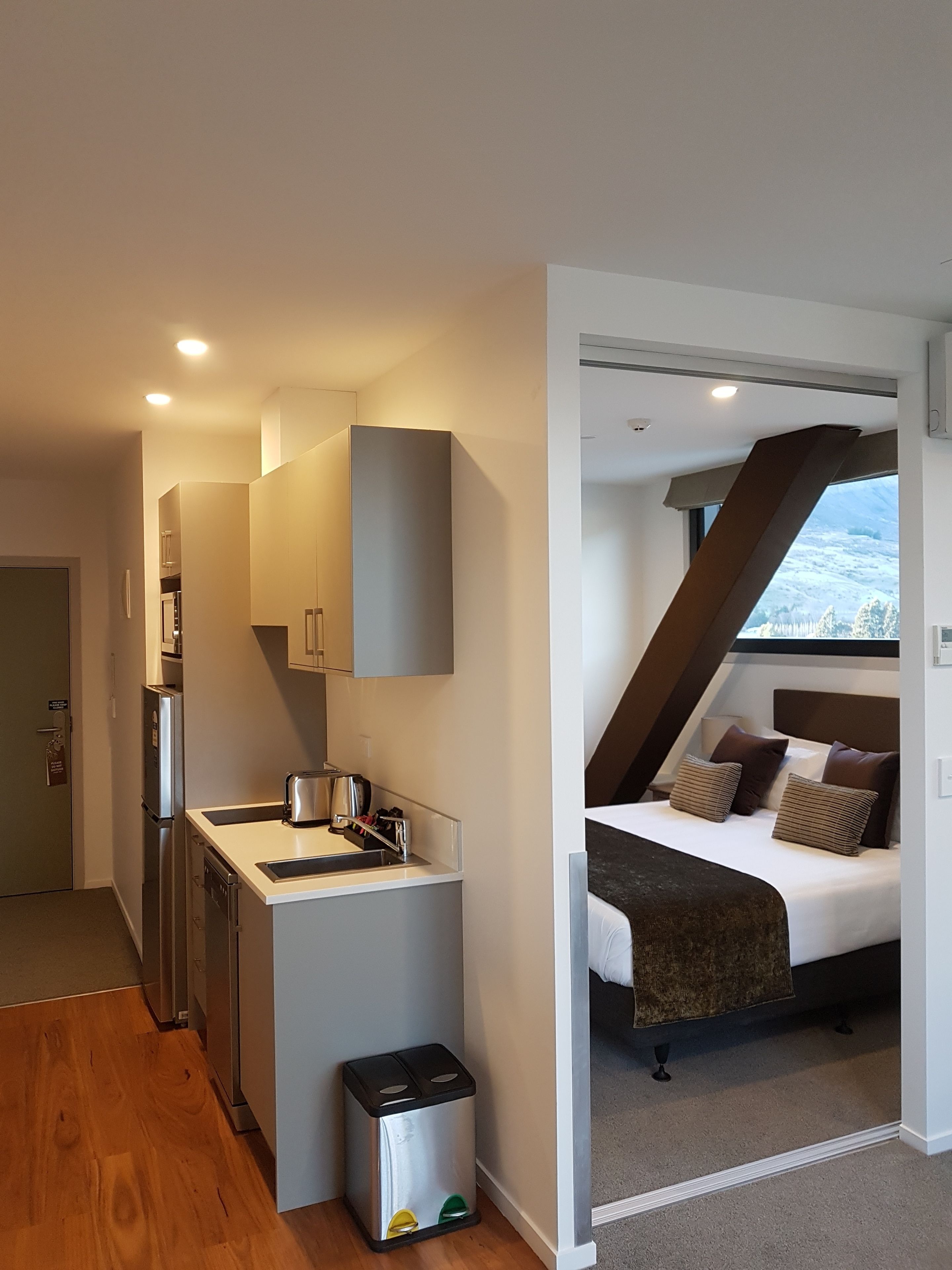 A compact hotel room with a kitchenette, a bed with a view, and a small trash bin.