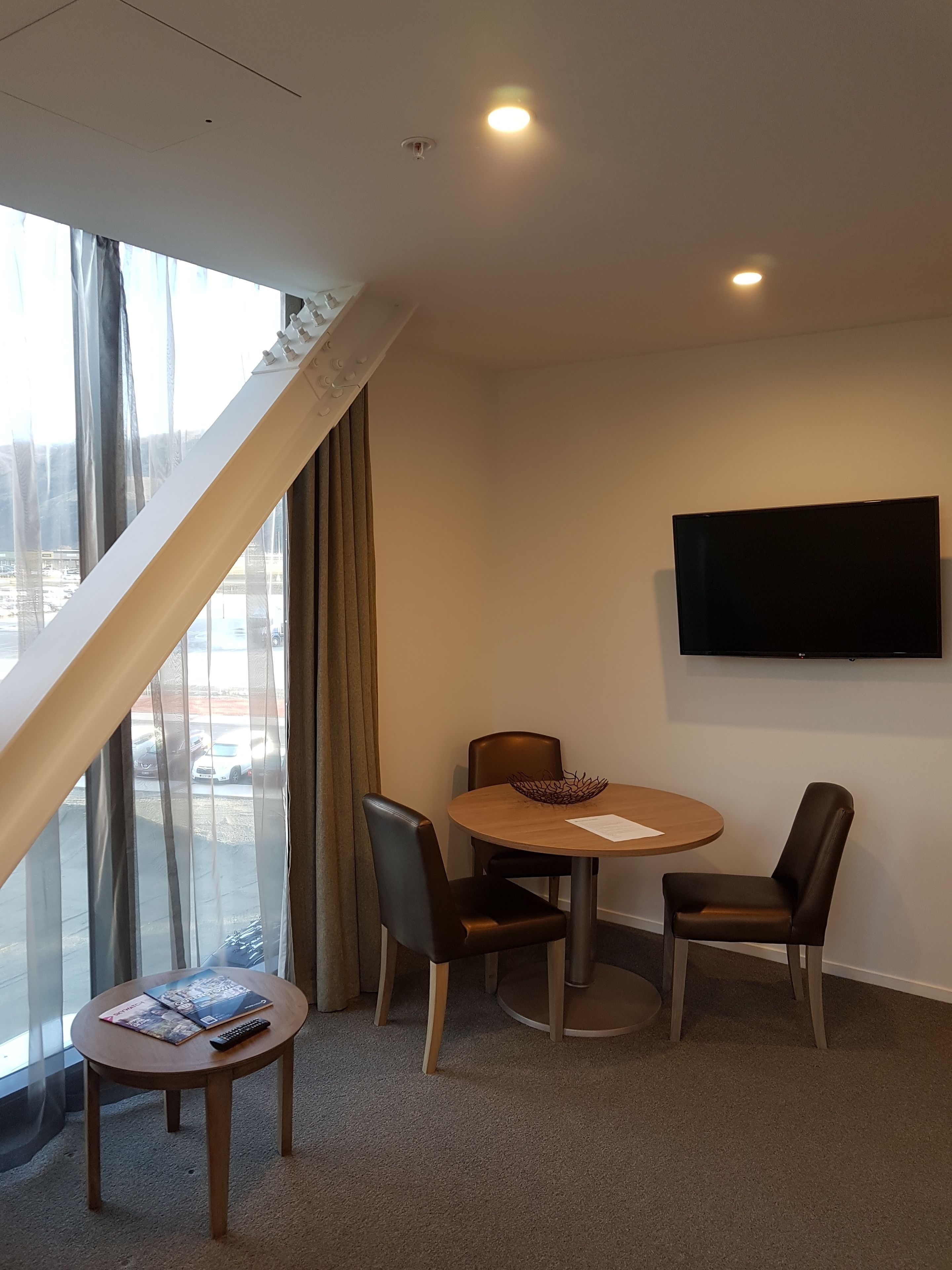 A hotel room with a large window, a round table with chairs, a flat-screen TV mounted on the wall, and a small table with a remote control.