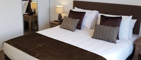 Un lit bien fait, avec une tête de lit marron, du linge de lit blanc et un plaid doré. Une table de chevet avec une lampe se trouve à côté du lit, et un tableau encadré est accroché au mur au-dessus du lit.