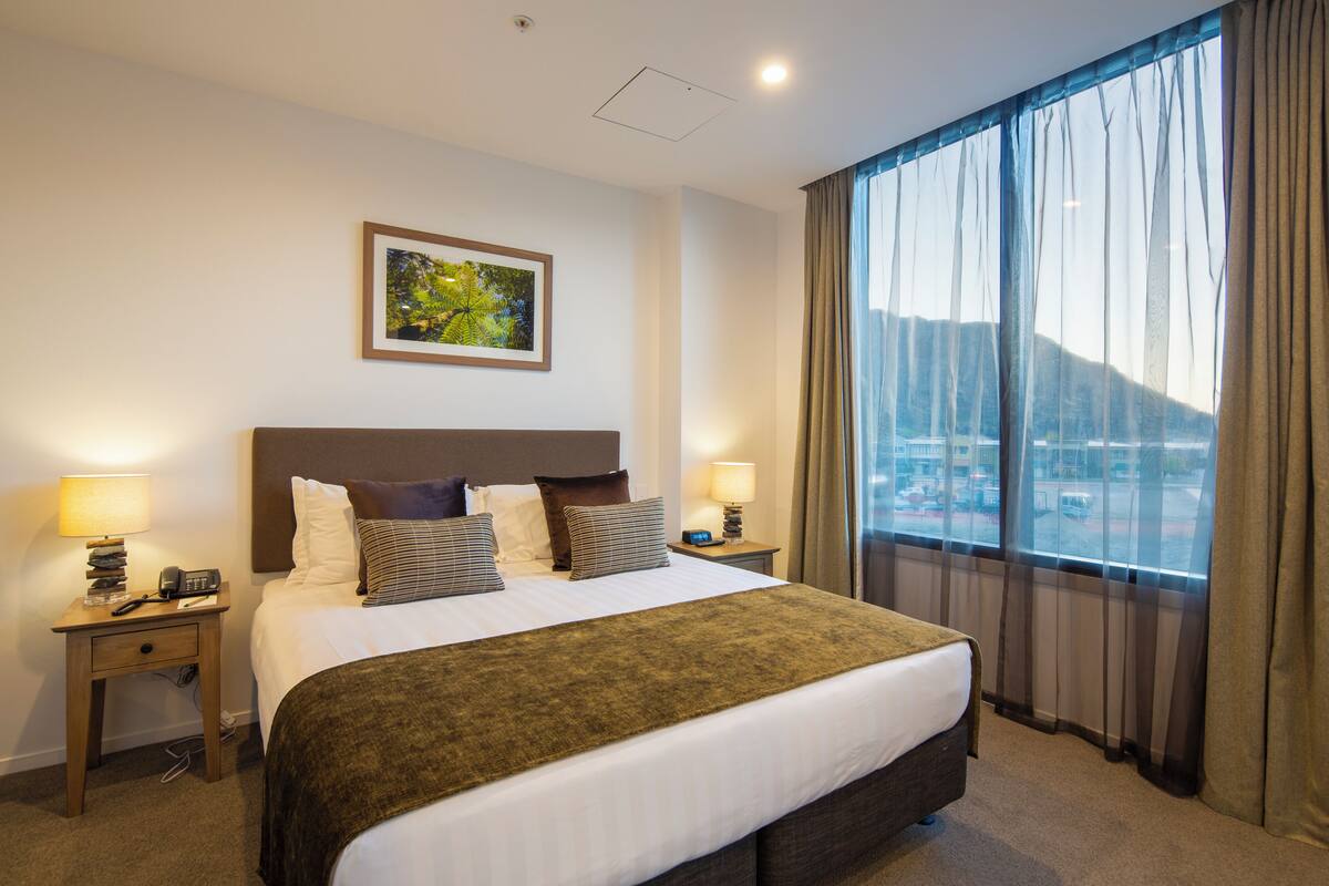 A hotel room with a large bed, two bedside tables, a nightstand, a framed picture on the wall, and a view of a cityscape through the window.