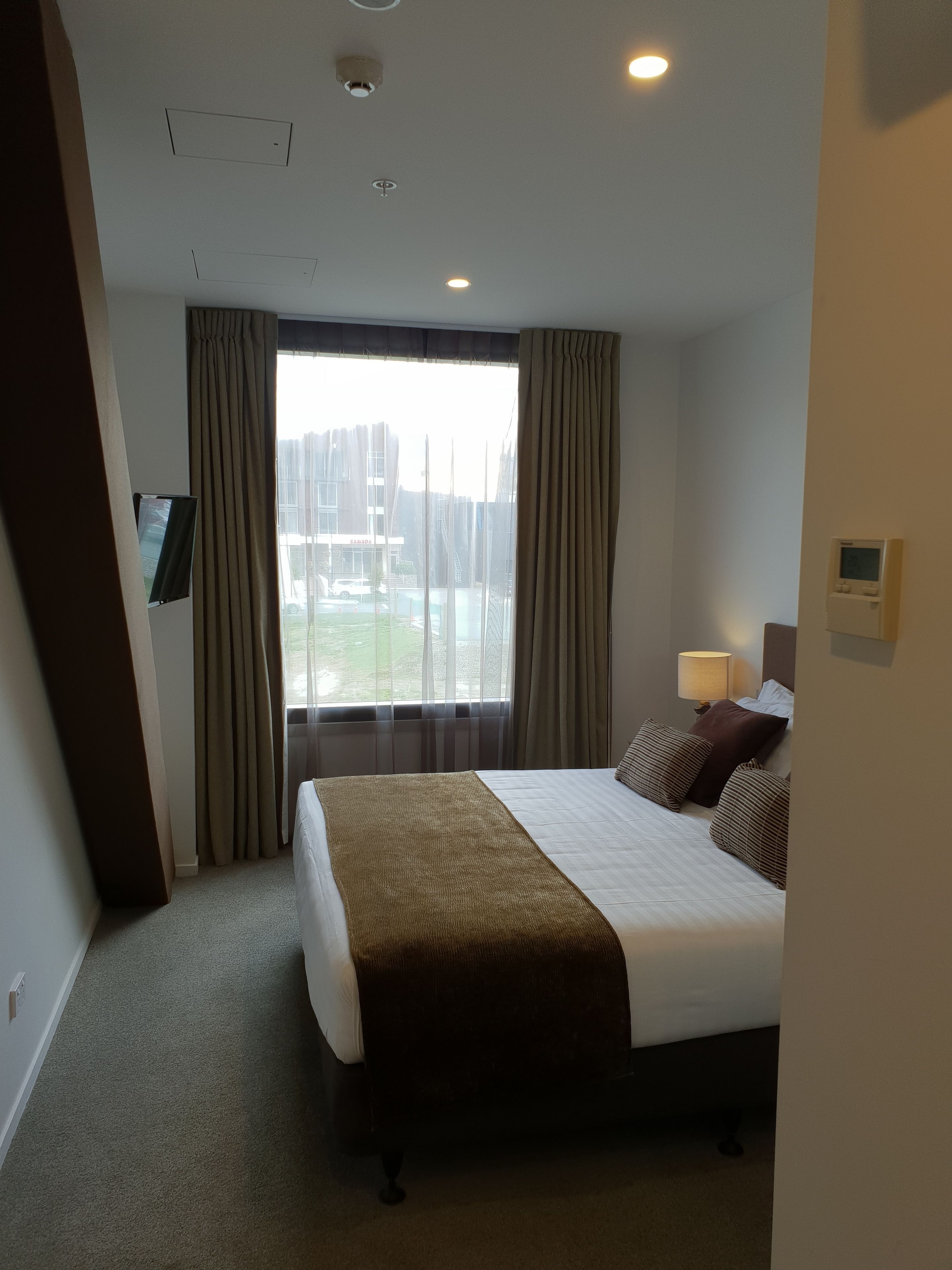 Ein Hotelzimmer mit einem großen Bett, einem an der Wand montierten Fernseher, einem Fenster mit Vorhängen und einem Thermostat.