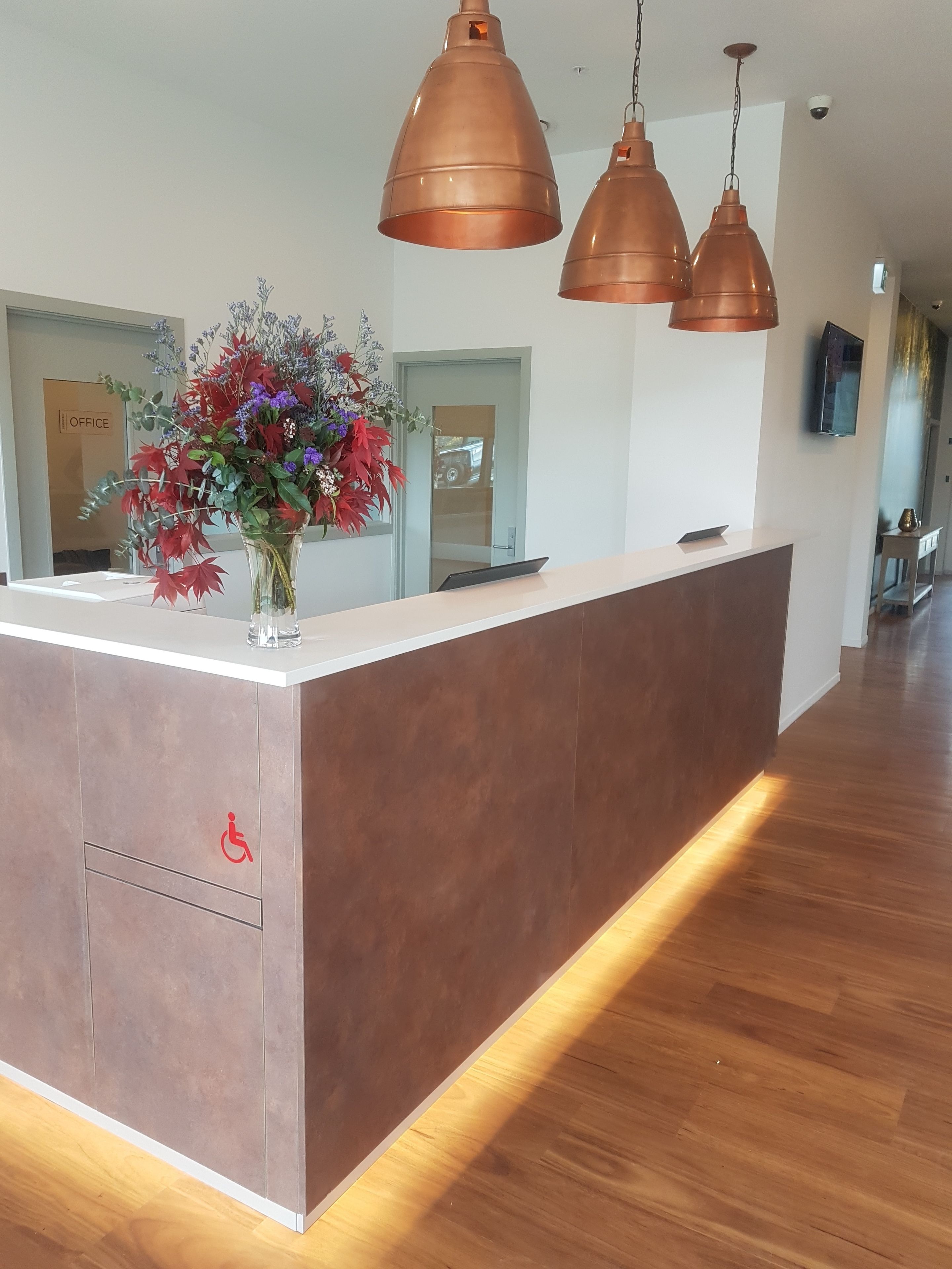 Un espace d’accueil avec un comptoir en bois, un arrangement floral et des luminaires suspendus en cuivre.