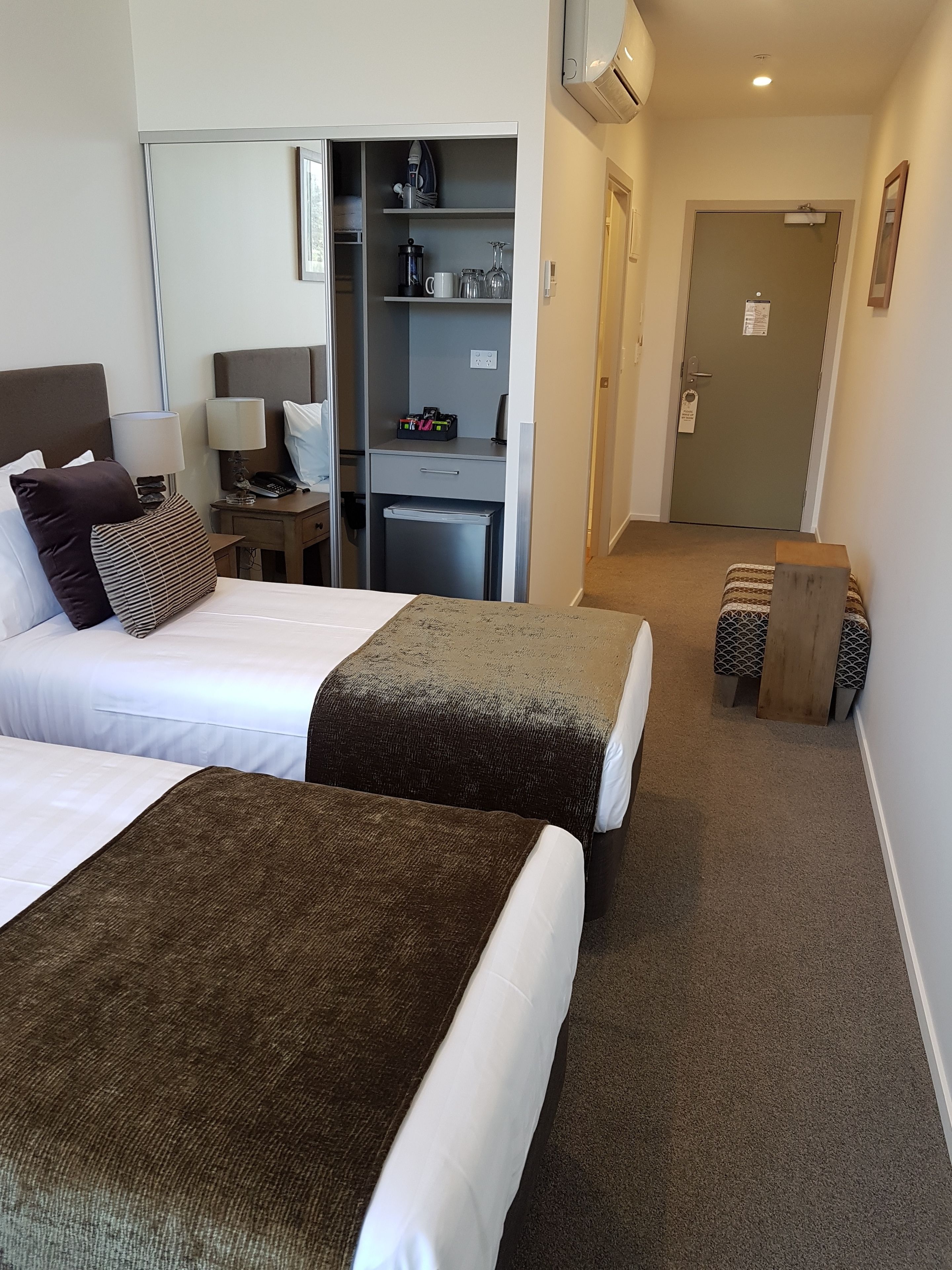 A hotel room with two beds, a desk with a lamp, a small kitchenette, and a door leading to a hallway.