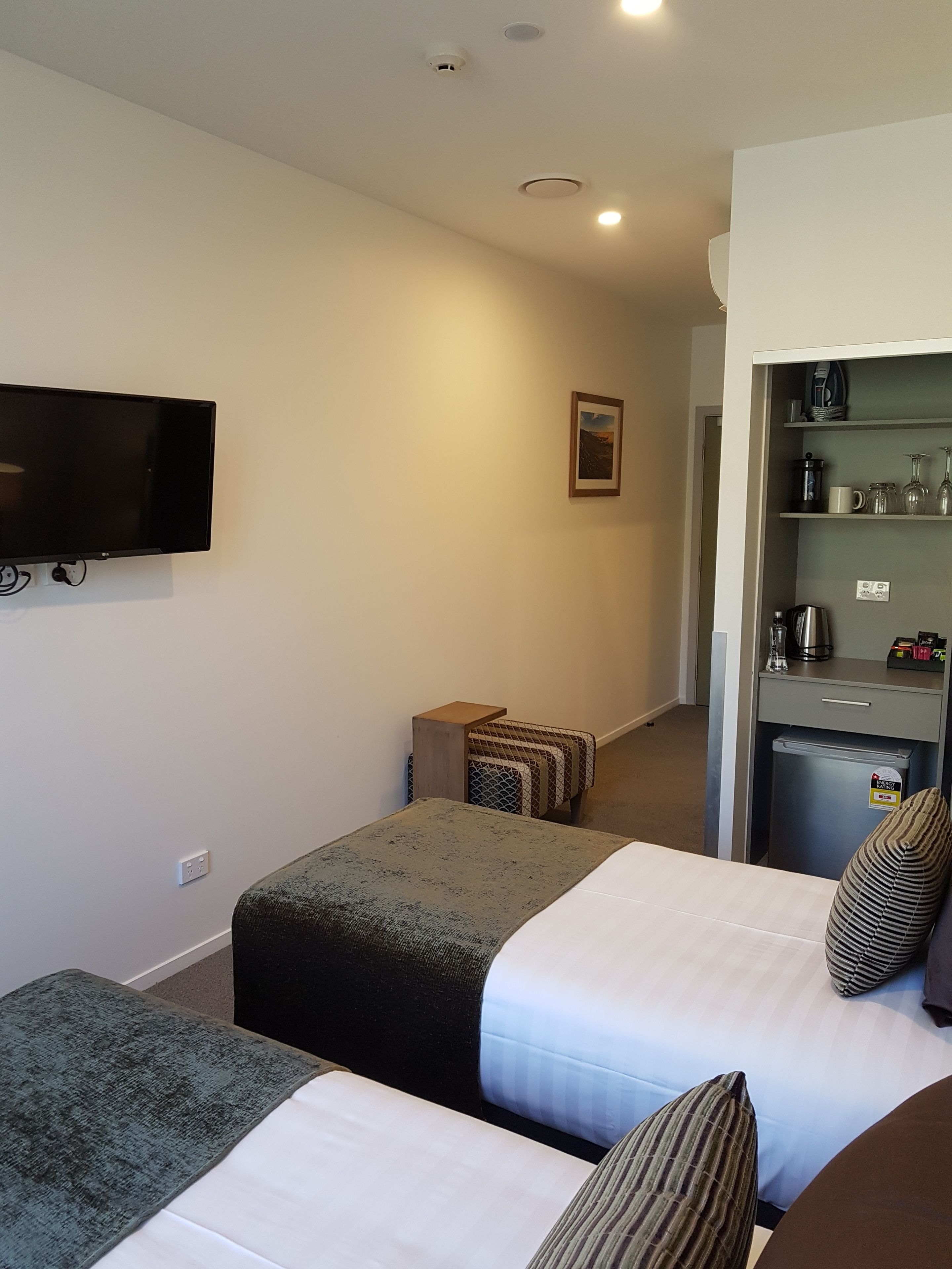 Habitación de hotel con dos camas, un televisor montado en la pared, una mesita auxiliar pequeña, una kitchenette con refrigerador y un cuadro enmarcado en la pared.