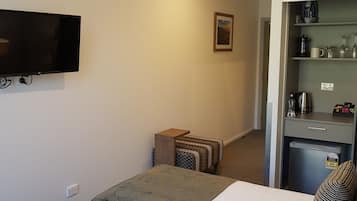 Habitación de hotel con dos camas, un televisor montado en la pared, una mesita auxiliar pequeña, una kitchenette con refrigerador y un cuadro enmarcado en la pared.