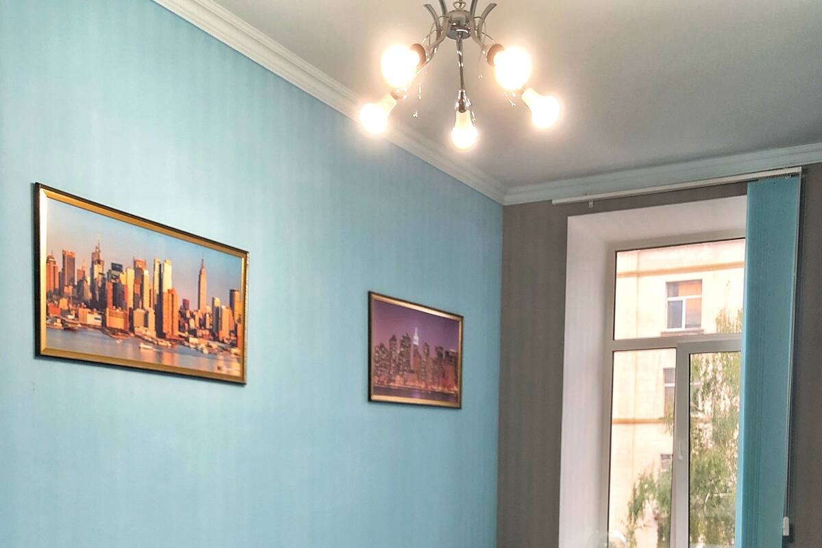 A bedroom with a brown leather sofa, a blue bedspread, and two framed cityscape paintings on the wall.