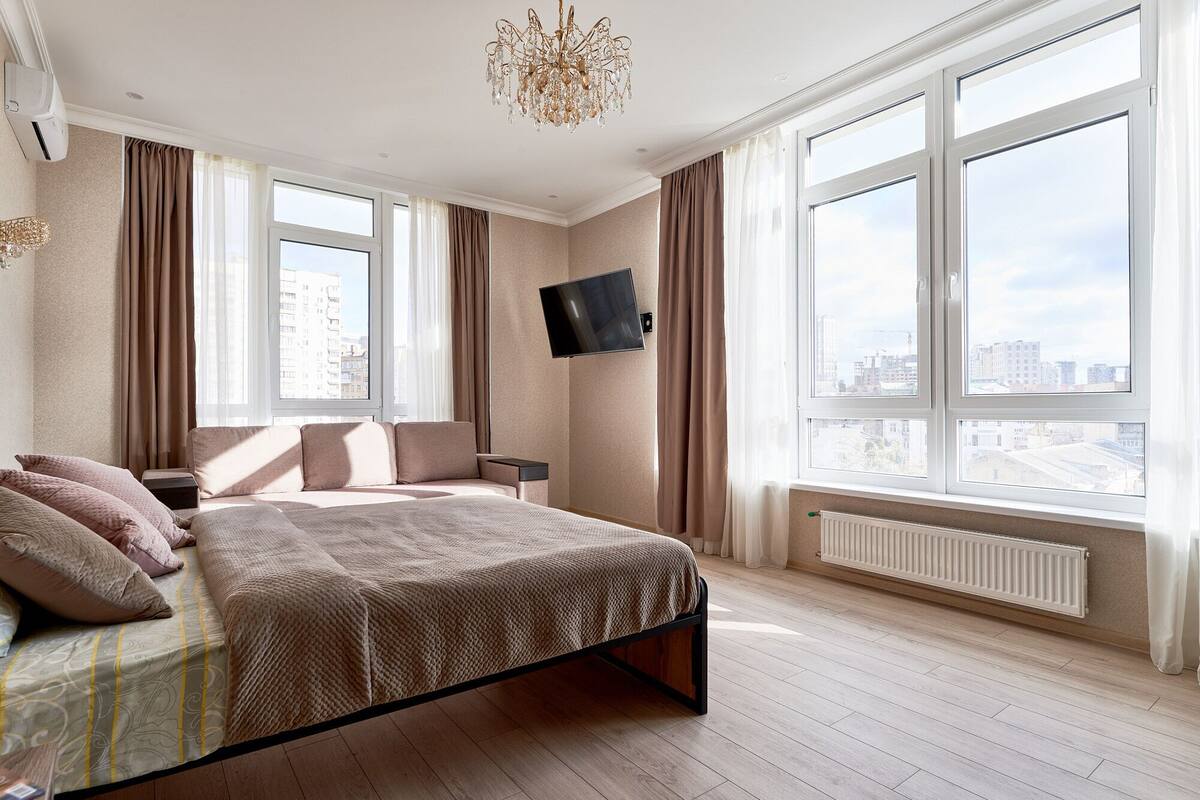 A modern hotel room with a large bed, a flat-screen TV mounted on the wall, and a city view through large windows.