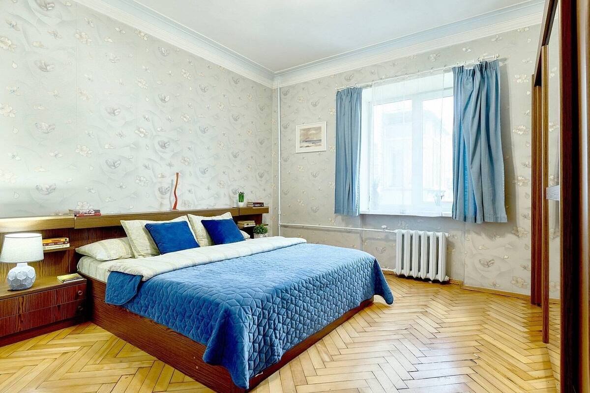 A bedroom with a wooden floor, a bed with a blue quilt, a nightstand with a lamp, and a window with curtains.