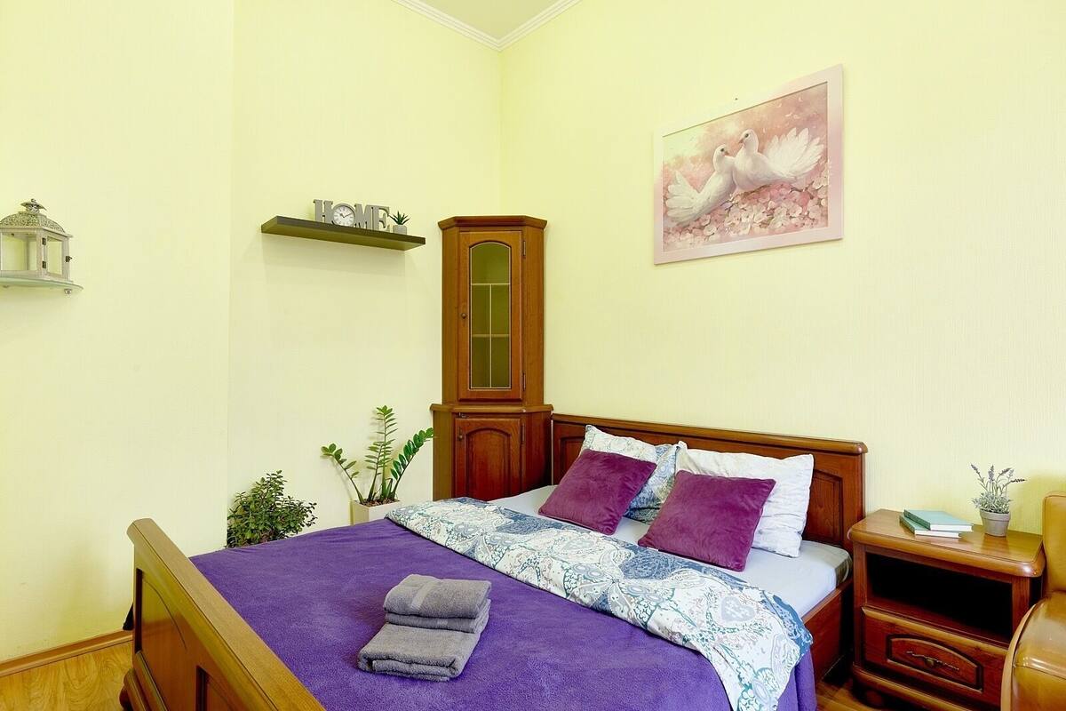 A neatly made bed with a purple blanket and pillows, a wooden headboard, a nightstand with a lamp, and a wall-mounted shelf with a 'HOME' sign.
