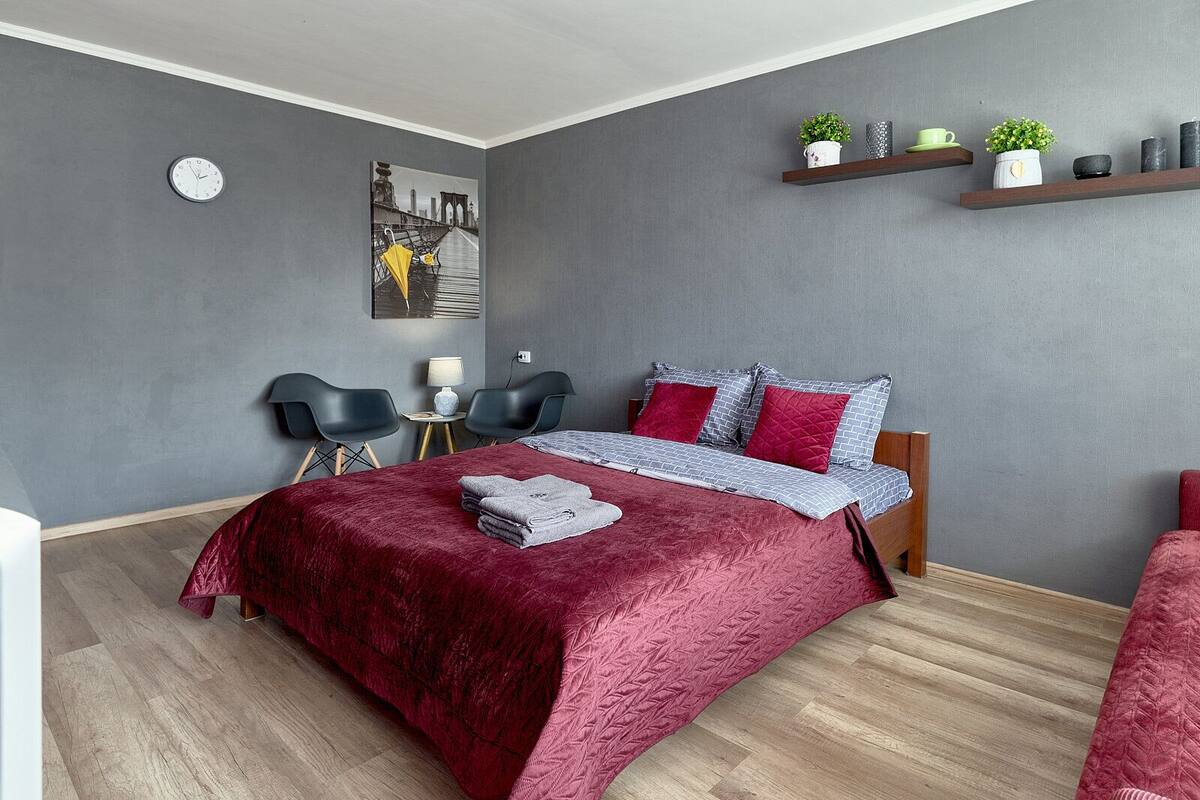 A modern bedroom with a large bed, grey walls, and a cityscape painting. There are two chairs, a small table, and a shelf with decorative items.