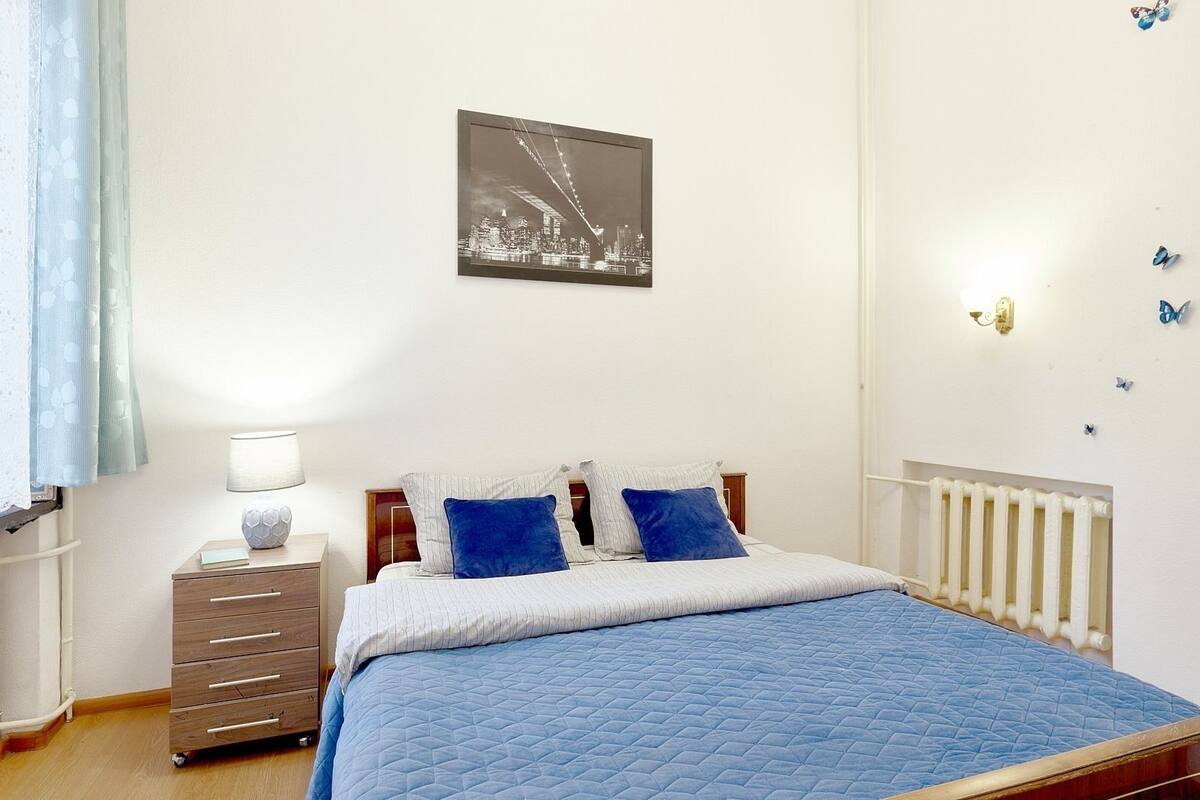 A bedroom with a bed, a nightstand, a lamp, a dresser, a radiator, and a cityscape picture on the wall.