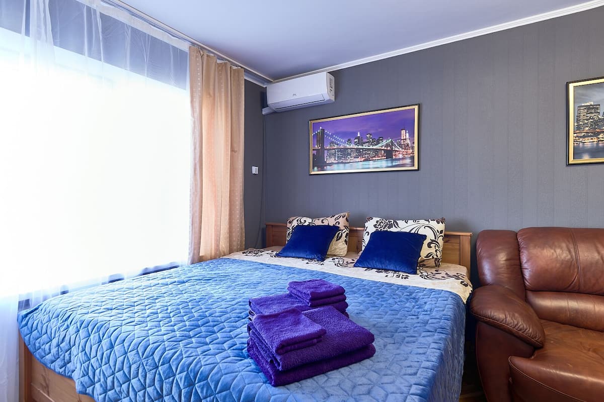 A hotel room with a bed, a brown leather armchair, a cityscape painting, and a window with curtains.