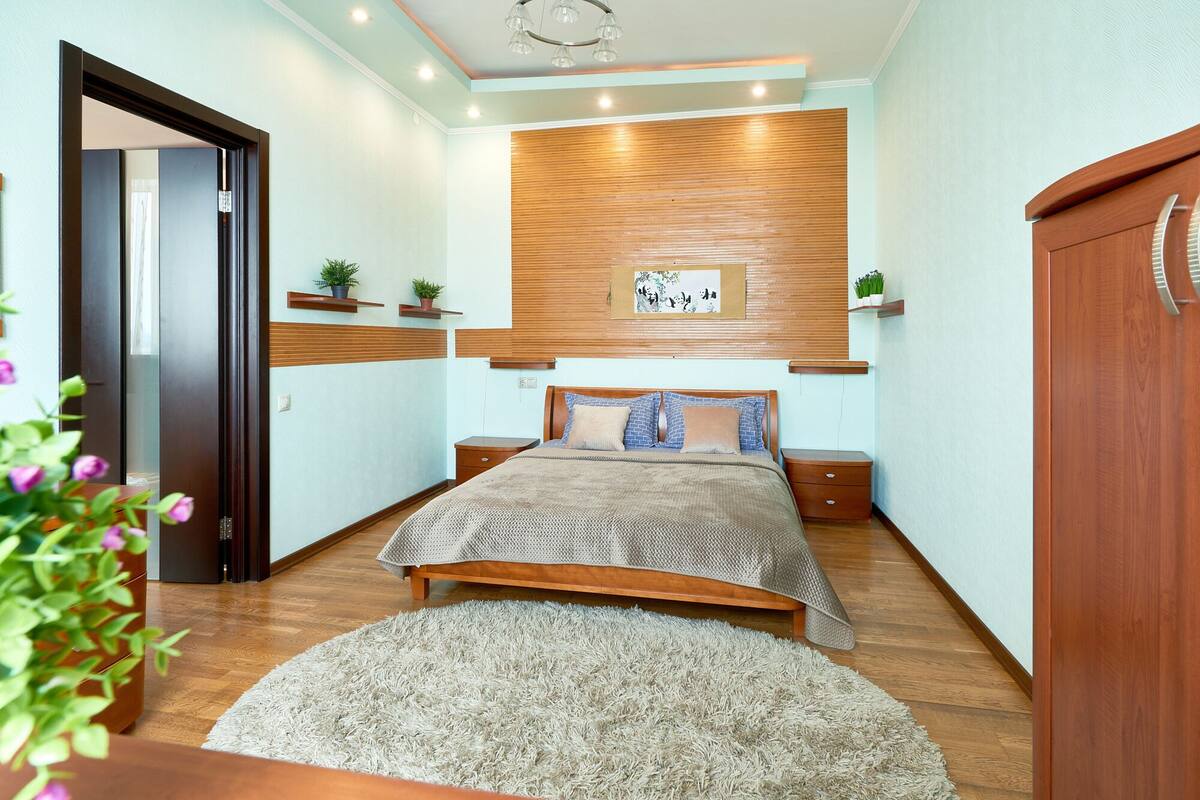 A bedroom with a wooden bed, bedside tables, a large rug, and wall-mounted shelves with plants.