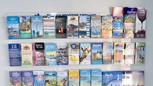 A wall of travel brochures and maps, including destinations like Swan River Cruises, Perth Zoo, and the Sunset Coast.
