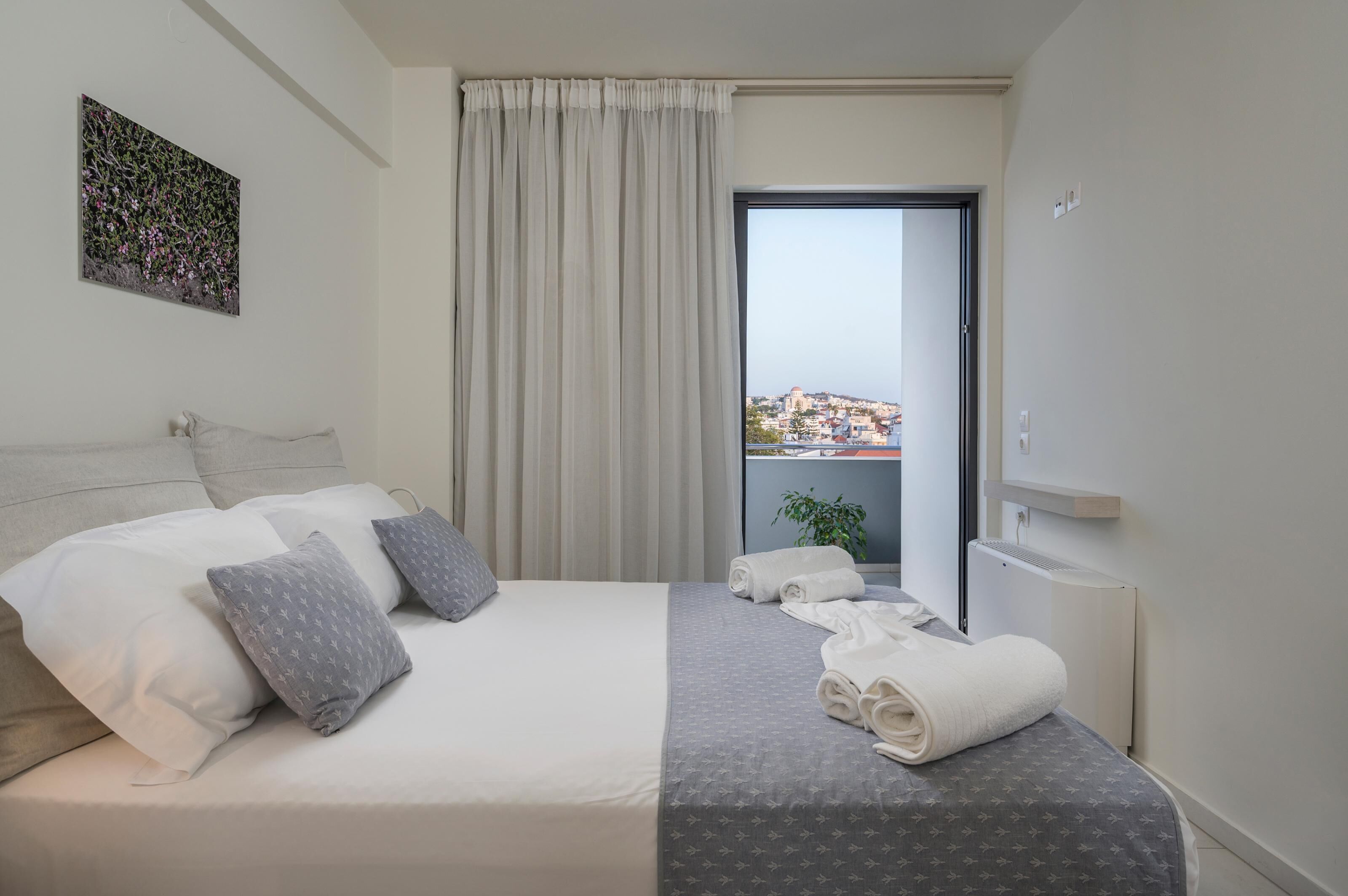 A hotel room with a bed, pillows, a view of a cityscape, a window with curtains, and a radiator.