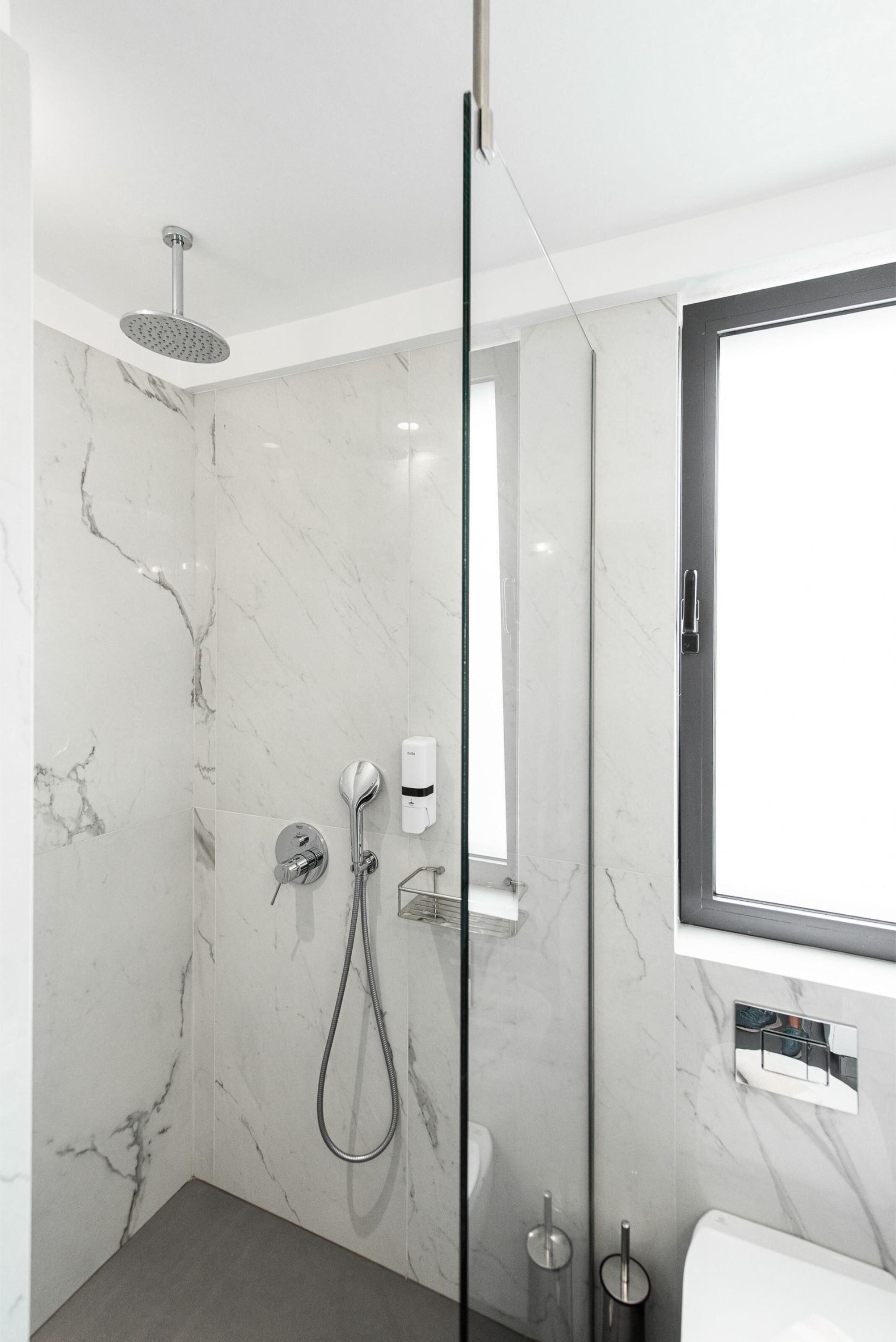 Un baño moderno con ducha envuelta en vidrio, paredes de mármol, inodoro y ventana.