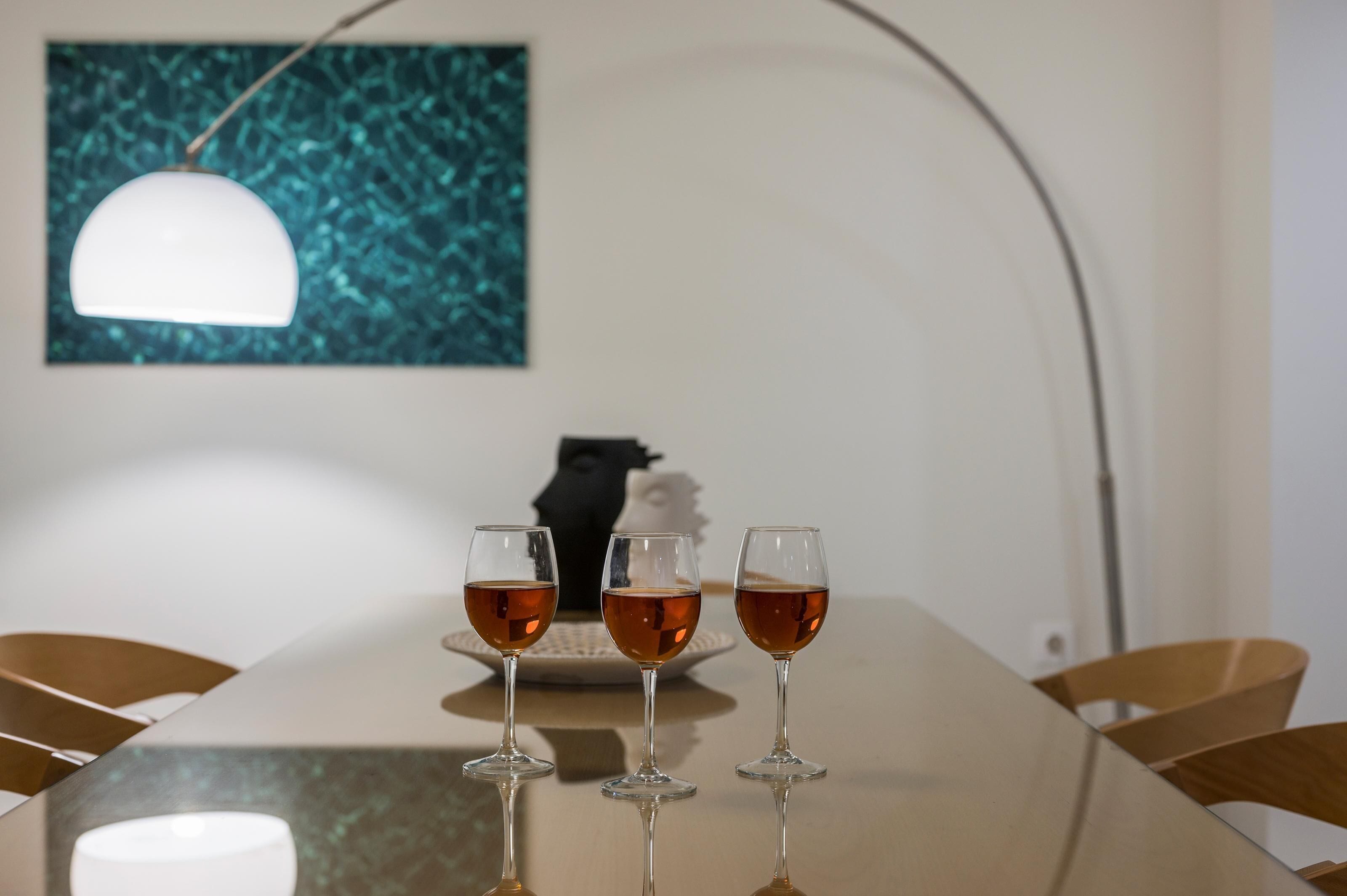 Una mesa de comedor con tres vasos de líquido ámbar, una lámpara colgante blanca y un cuadro abstracto color turquesa en la pared.