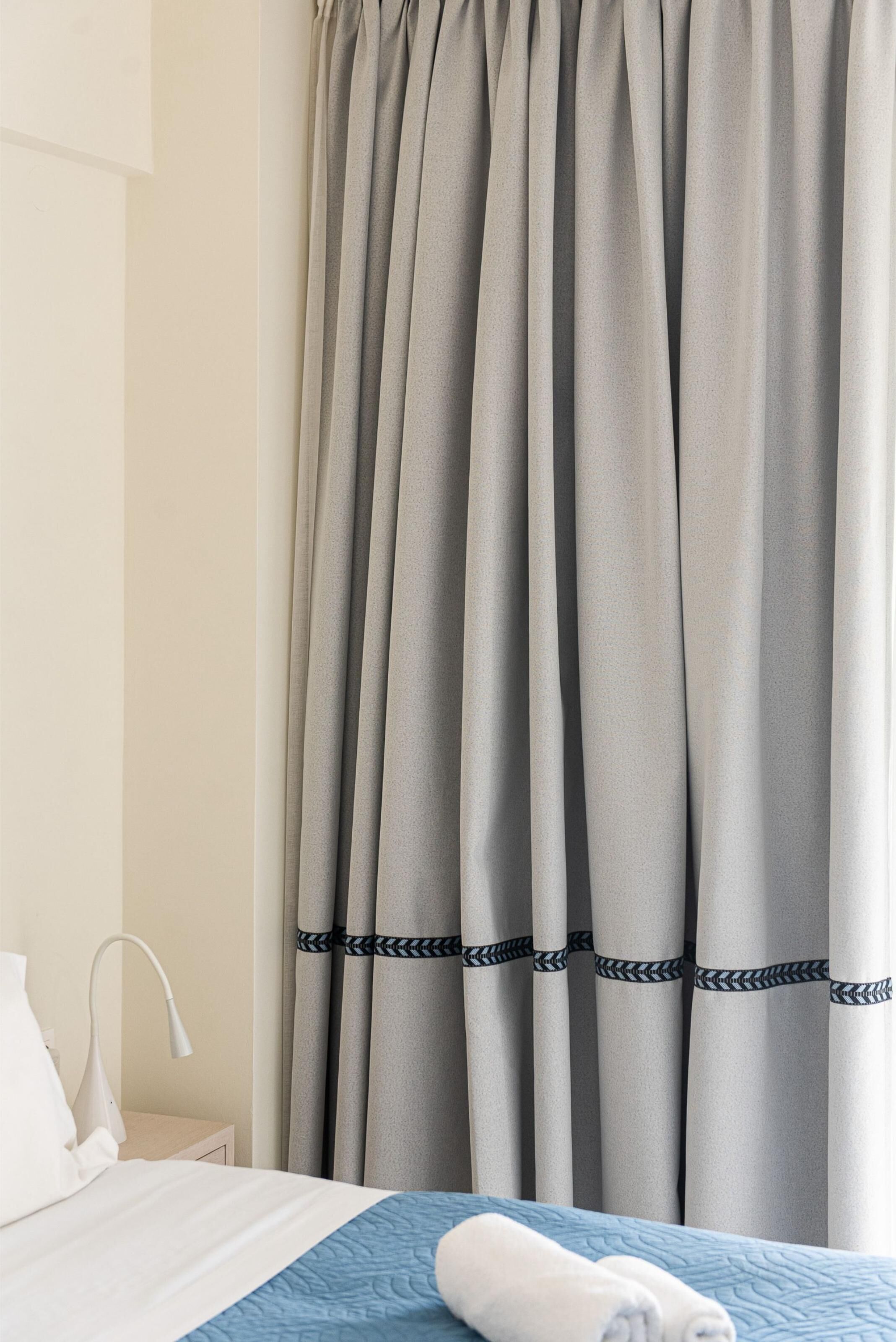 A hotel room with a bed, a nightstand, a lamp, and grey curtains with a black band.