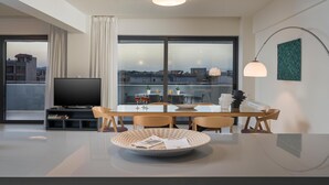 A modern living room with a large TV, dining table, and chairs. There is a large window with a view of the city. - Spring Apartments (Chania)