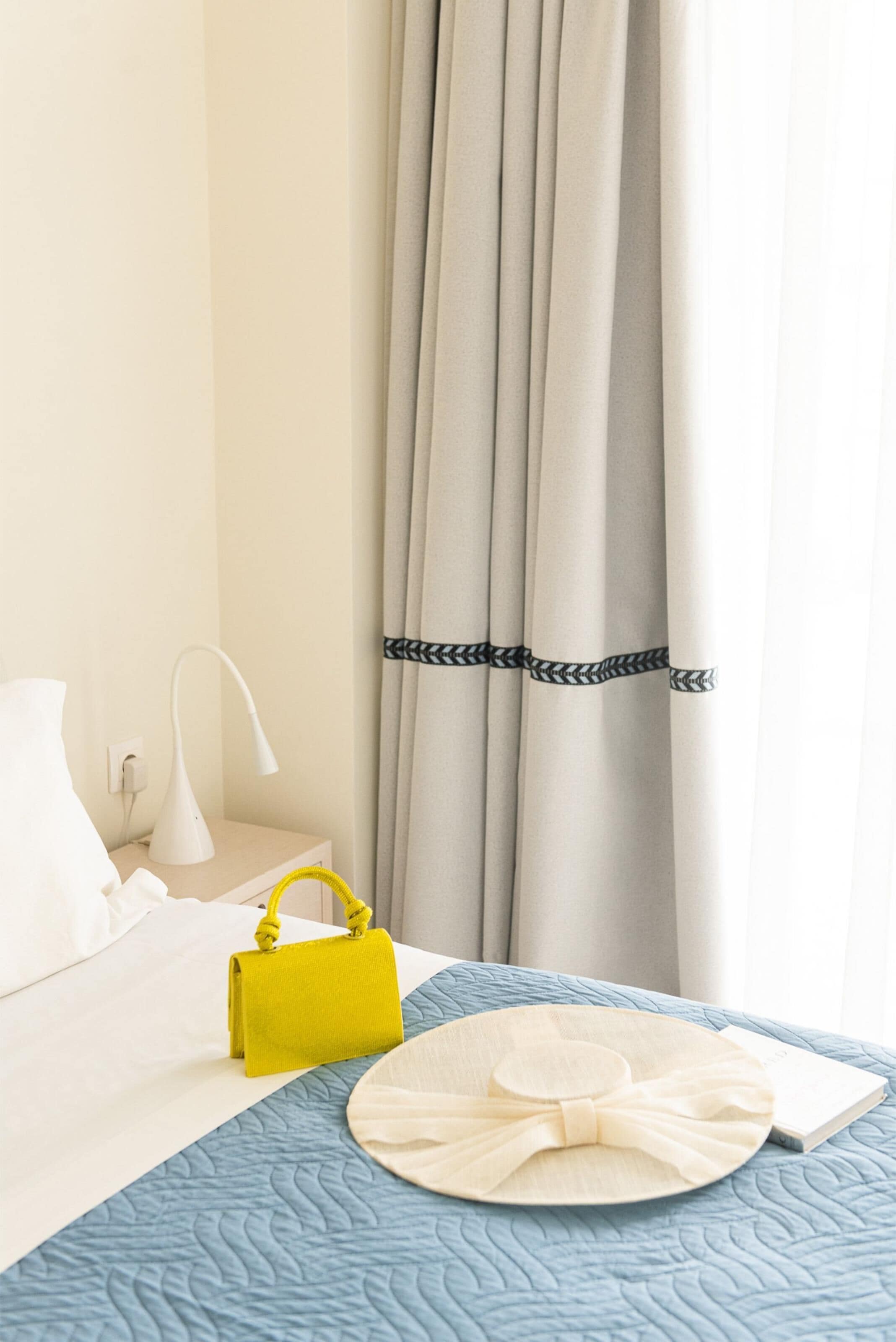 Una cama bien hecha con colcha azul, almohada blanca, lámpara blanca, bolso amarillo y un libro en la mesita de noche.